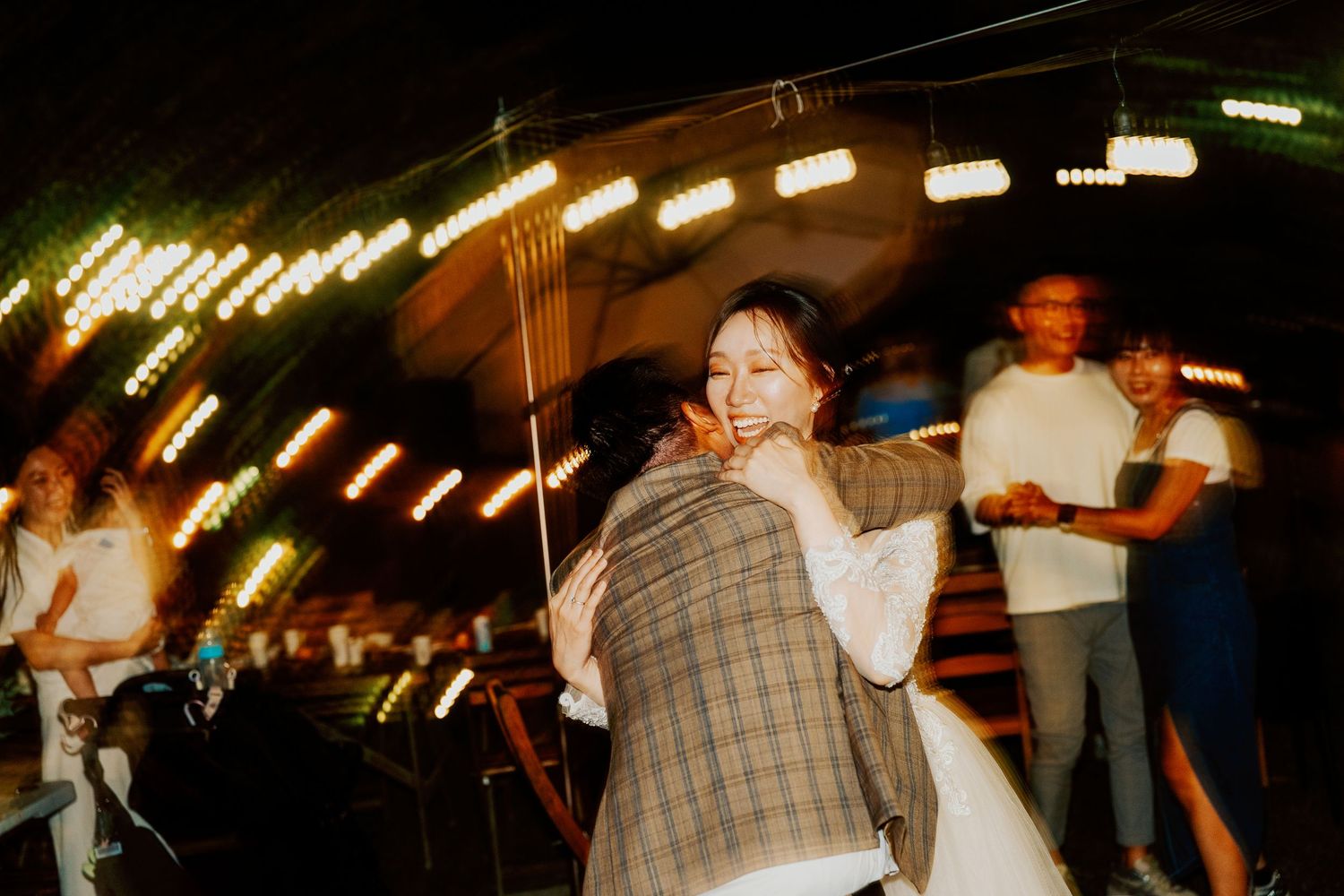 People dancing and enjoying outdoor festivities in a tent with festive string lights at night.