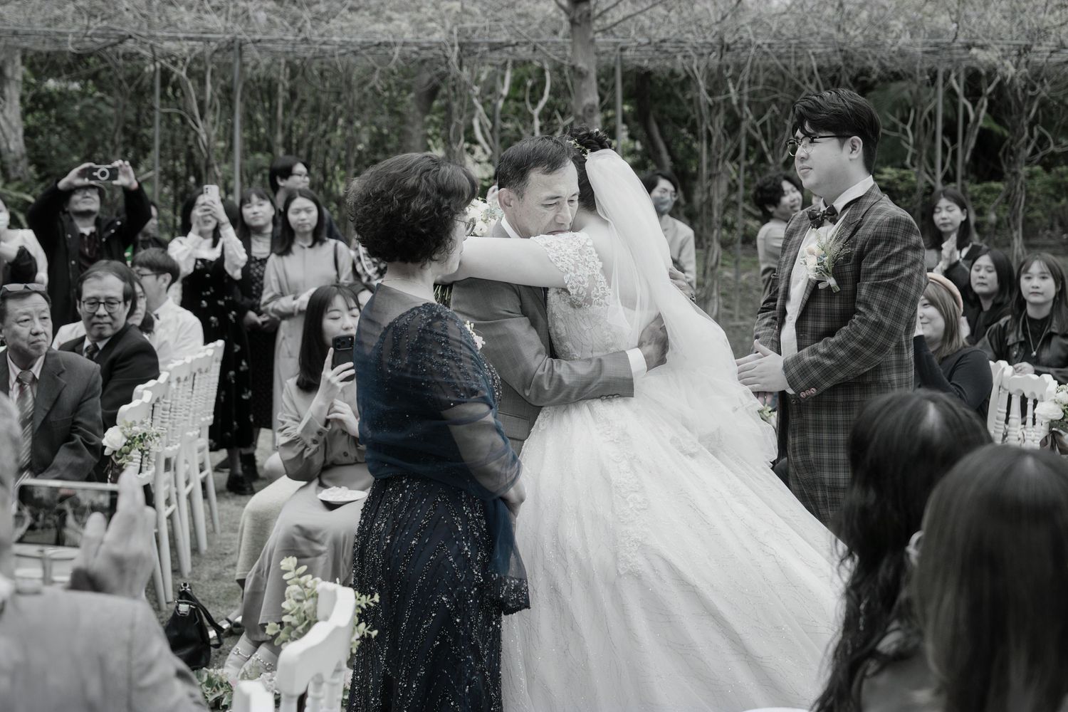 A vintage black and white wedding ceremony photo showing guests gathered outdoors as a bride in a full white gown embraces someone.