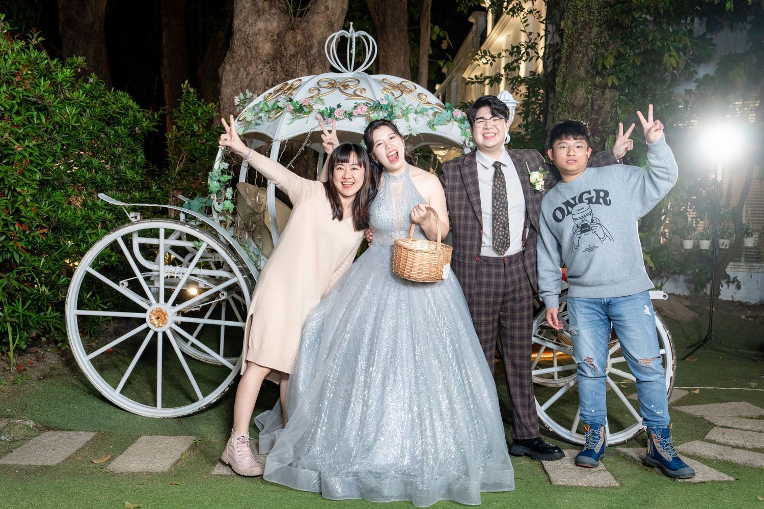 People gathered outdoors at a nighttime event wearing formal attire and posing near a decorative carriage wheel.