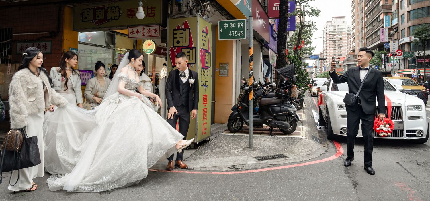 Wedding photographer captures panoramic shot of bridal party on city street corner with parked cars and buildings.
