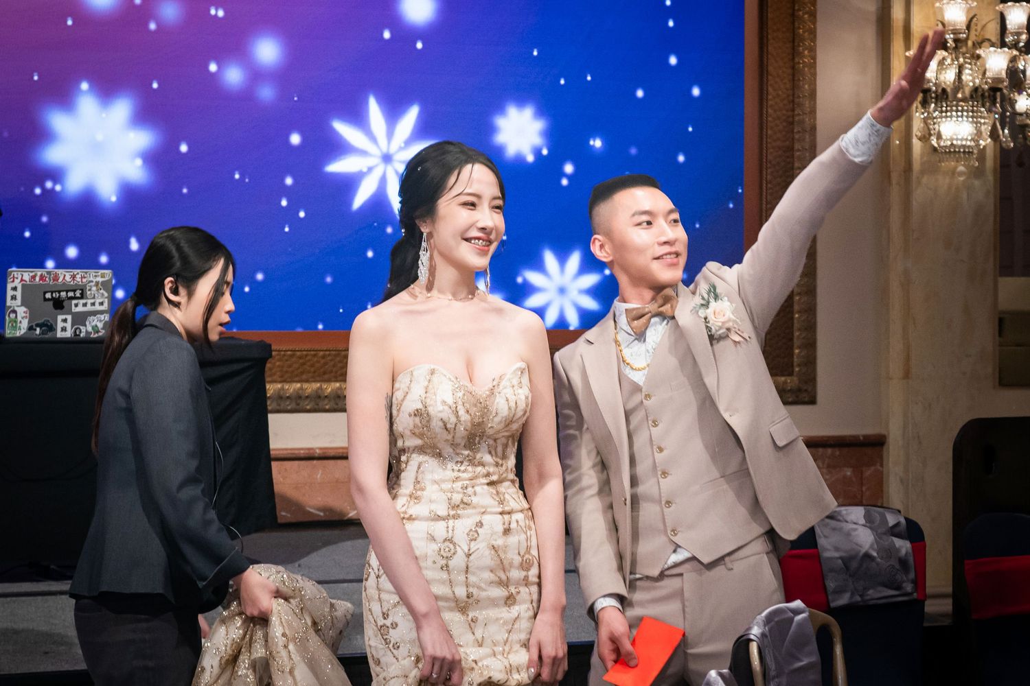 A couple celebrates at an elegant event with a blue starry backdrop while another person assists nearby.