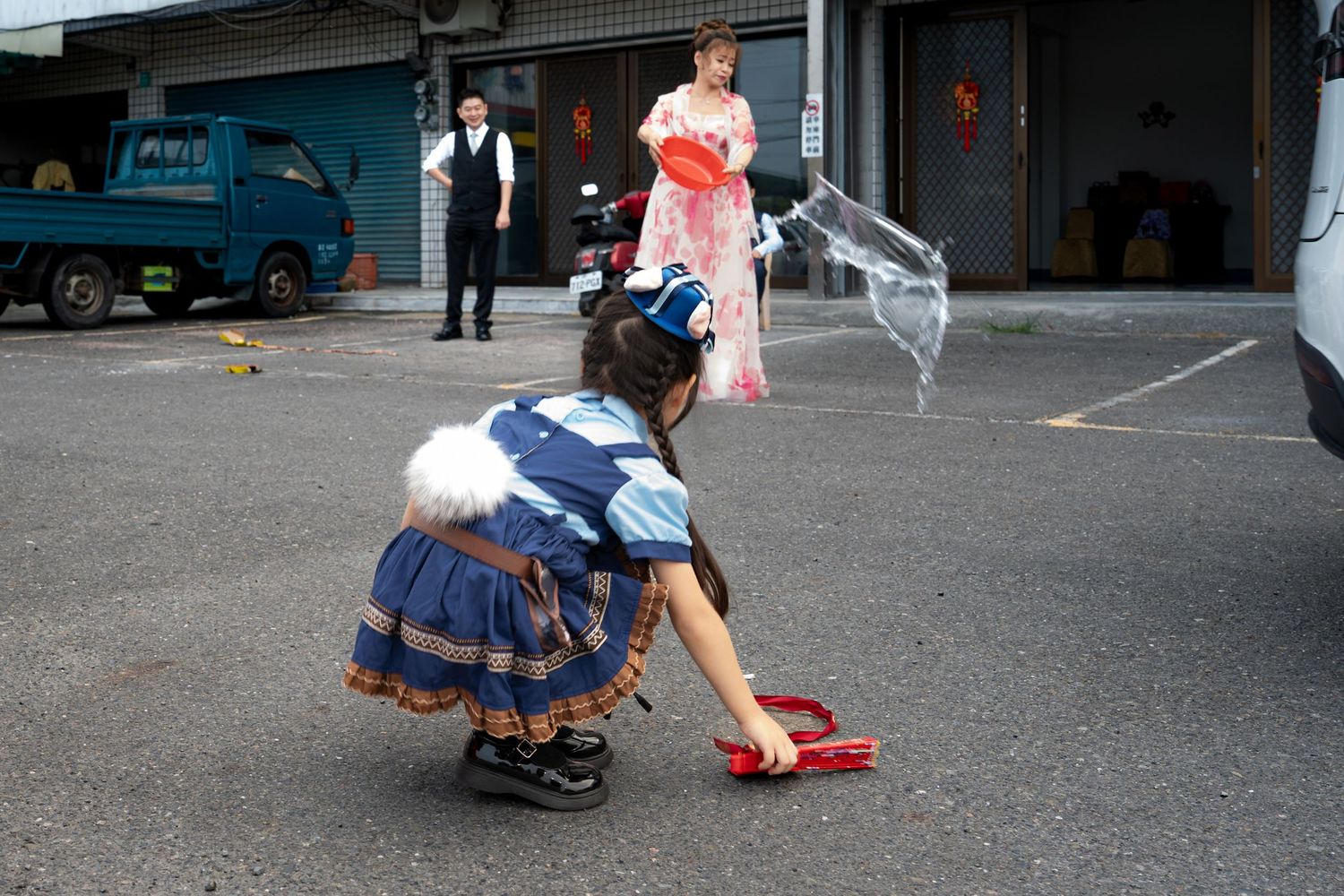 Child in blue costume picks up red shoes in parking lot, with woman in pink kimono and man in suit behind her.