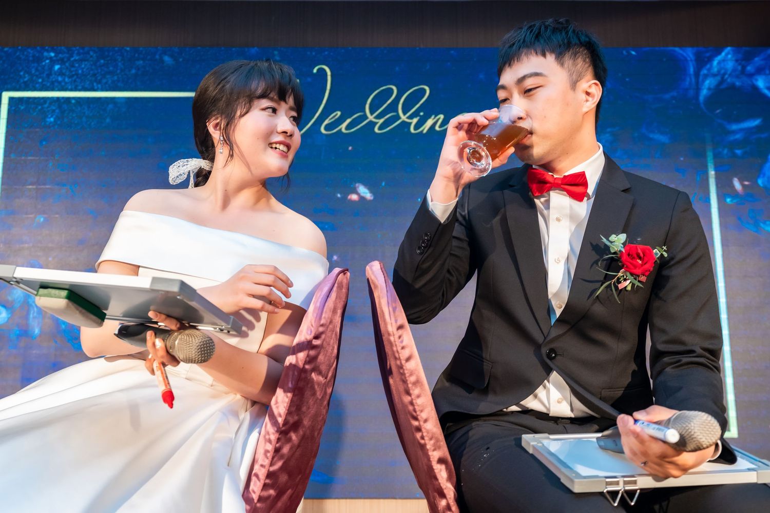 A bride in white dress and groom in tuxedo participate in a drinking game at their reception.