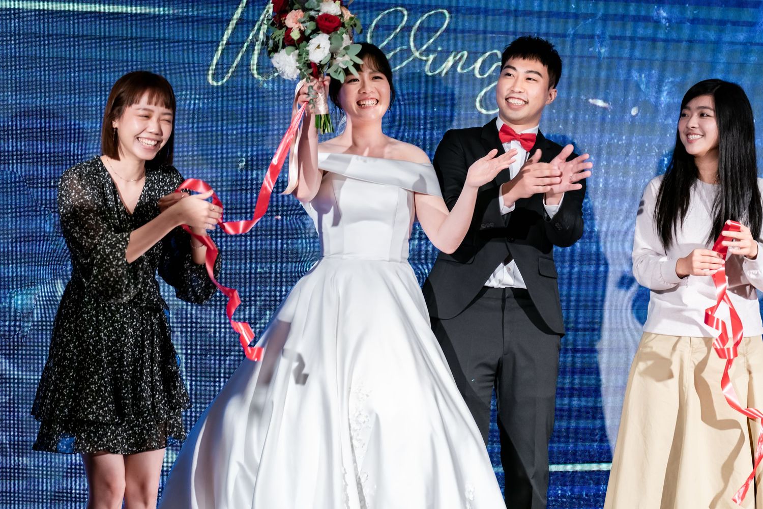 Wedding party celebrates with red ribbons during reception against blue backdrop with neon sign.