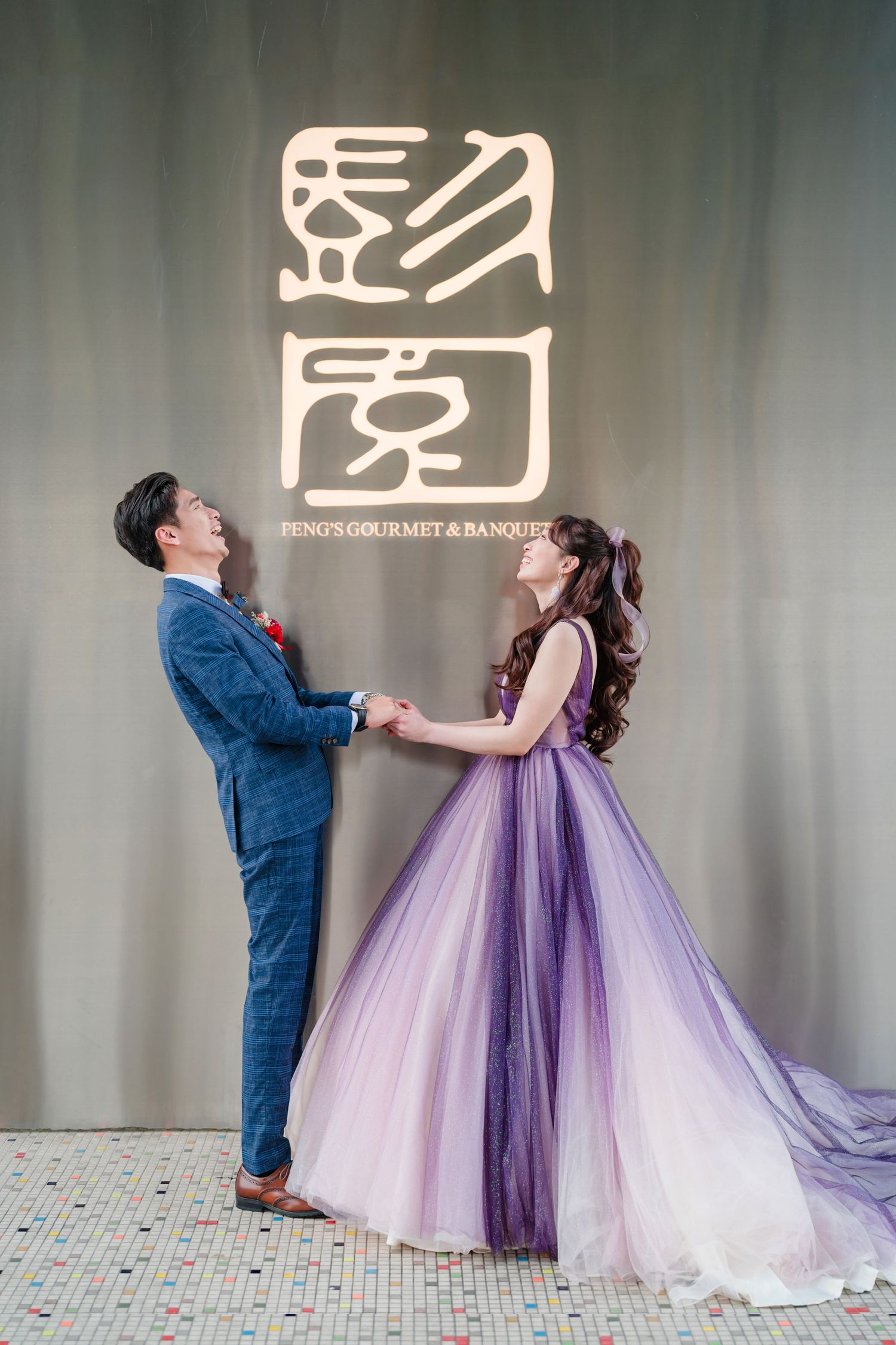 A couple poses together at a wedding venue, with the bride wearing a flowing purple ombre gown against a grey backdrop.