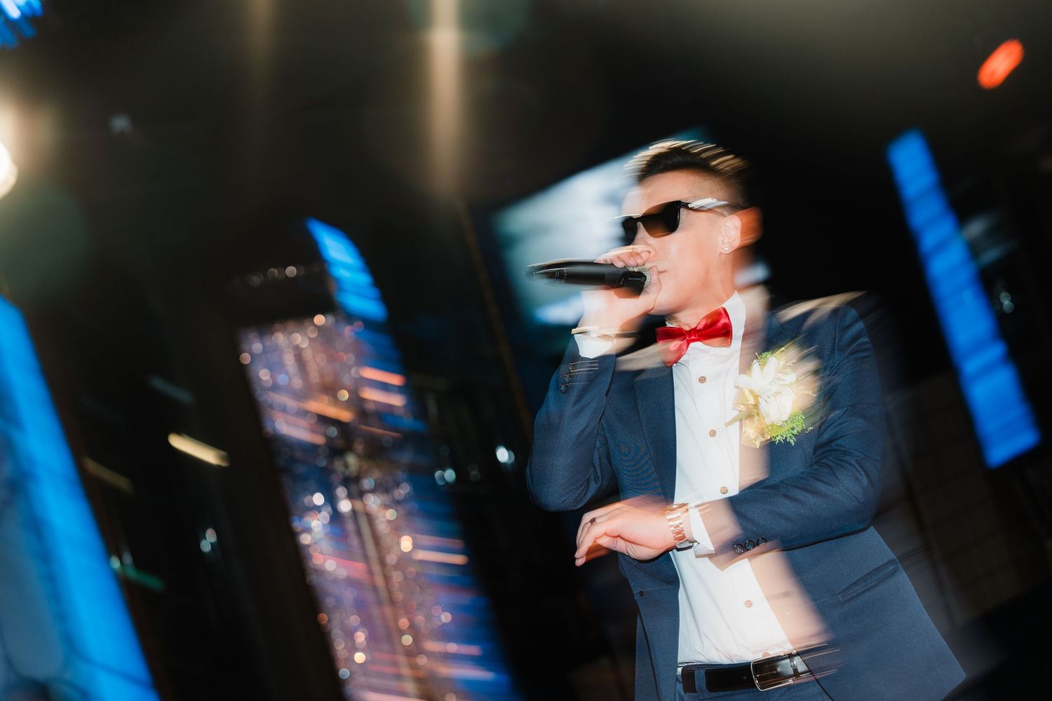 A performer in a suit and bow tie sings on stage with blue lighting and motion blur effects during a live show.