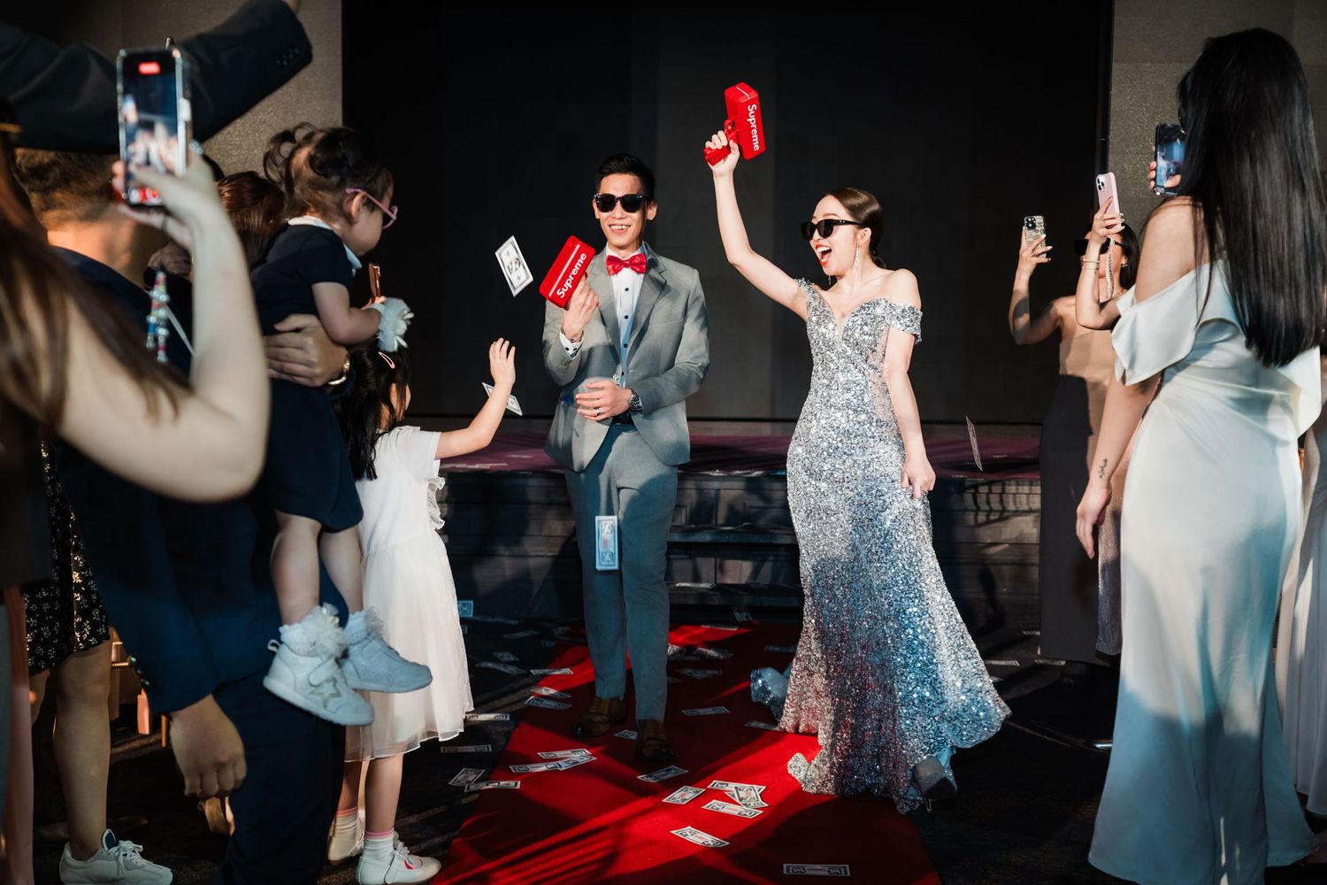Guests on a red carpet dance floor hold up phones during a festive celebration at a darkly lit evening event.