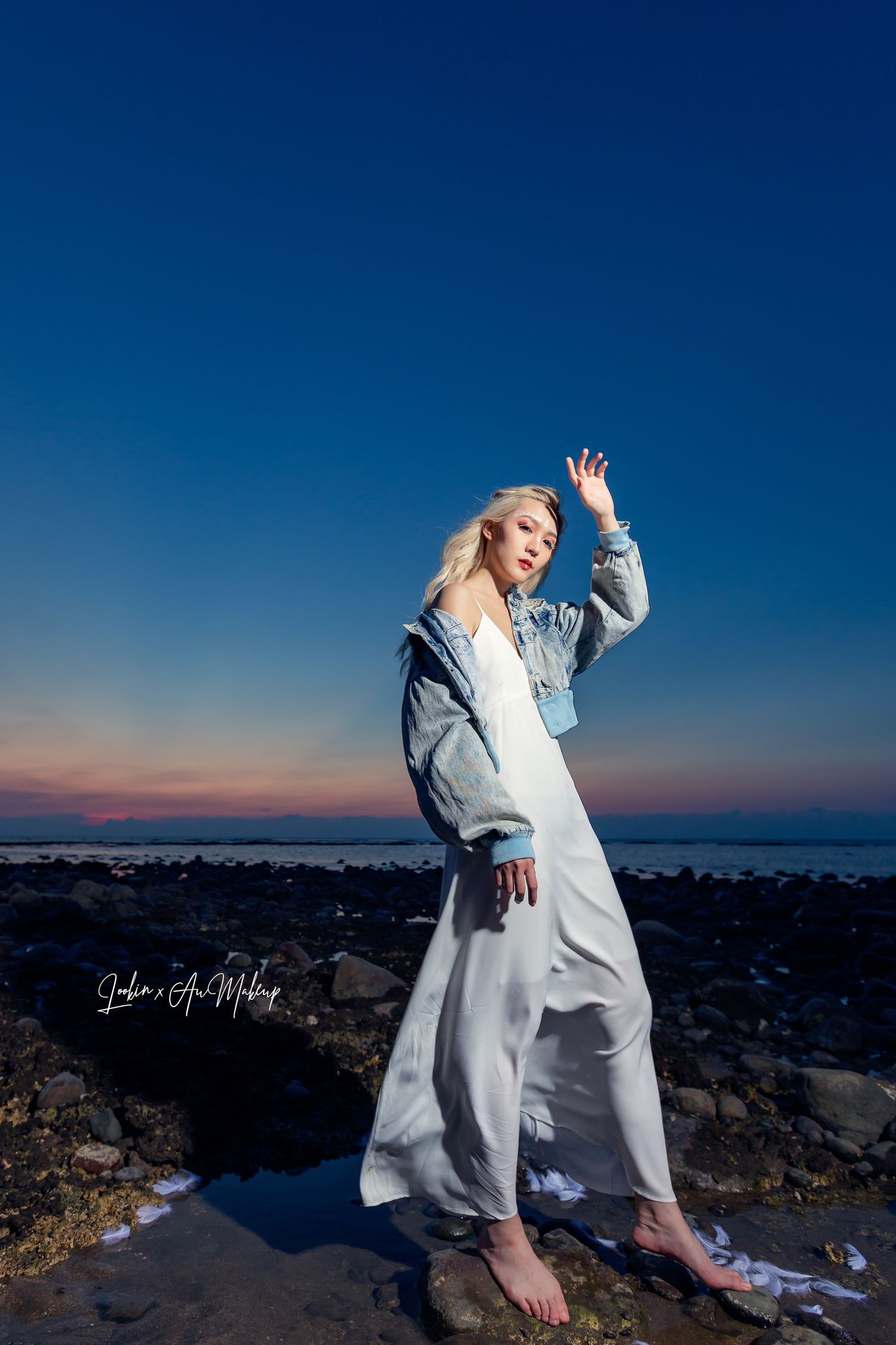 A person in a white dress and denim jacket poses barefoot on rocky coastal terrain at sunset against a deep blue sky.