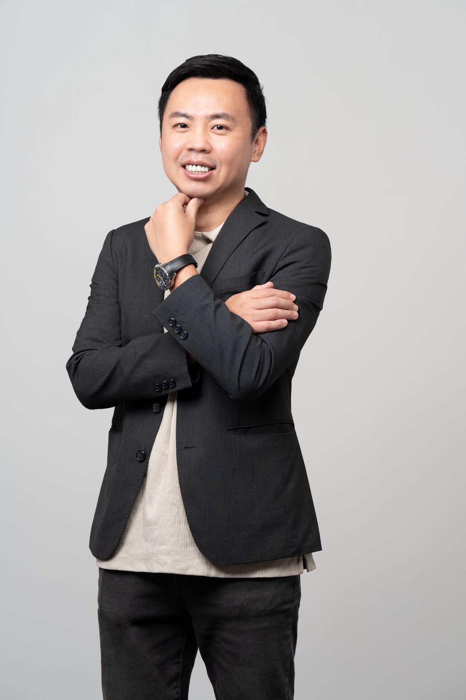 Professional portrait of a person in a black blazer and casual attire posing with hand on chin against gray background.