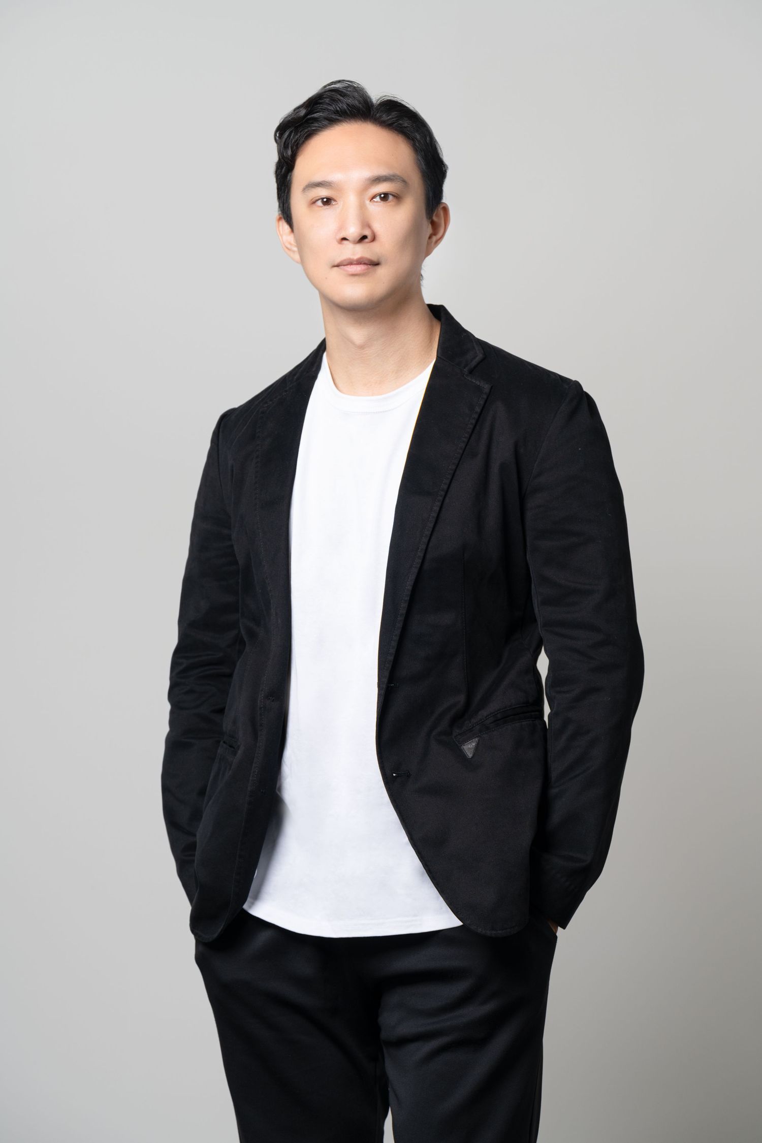 A professional studio portrait of a person wearing a black blazer and white t-shirt against a gray background.