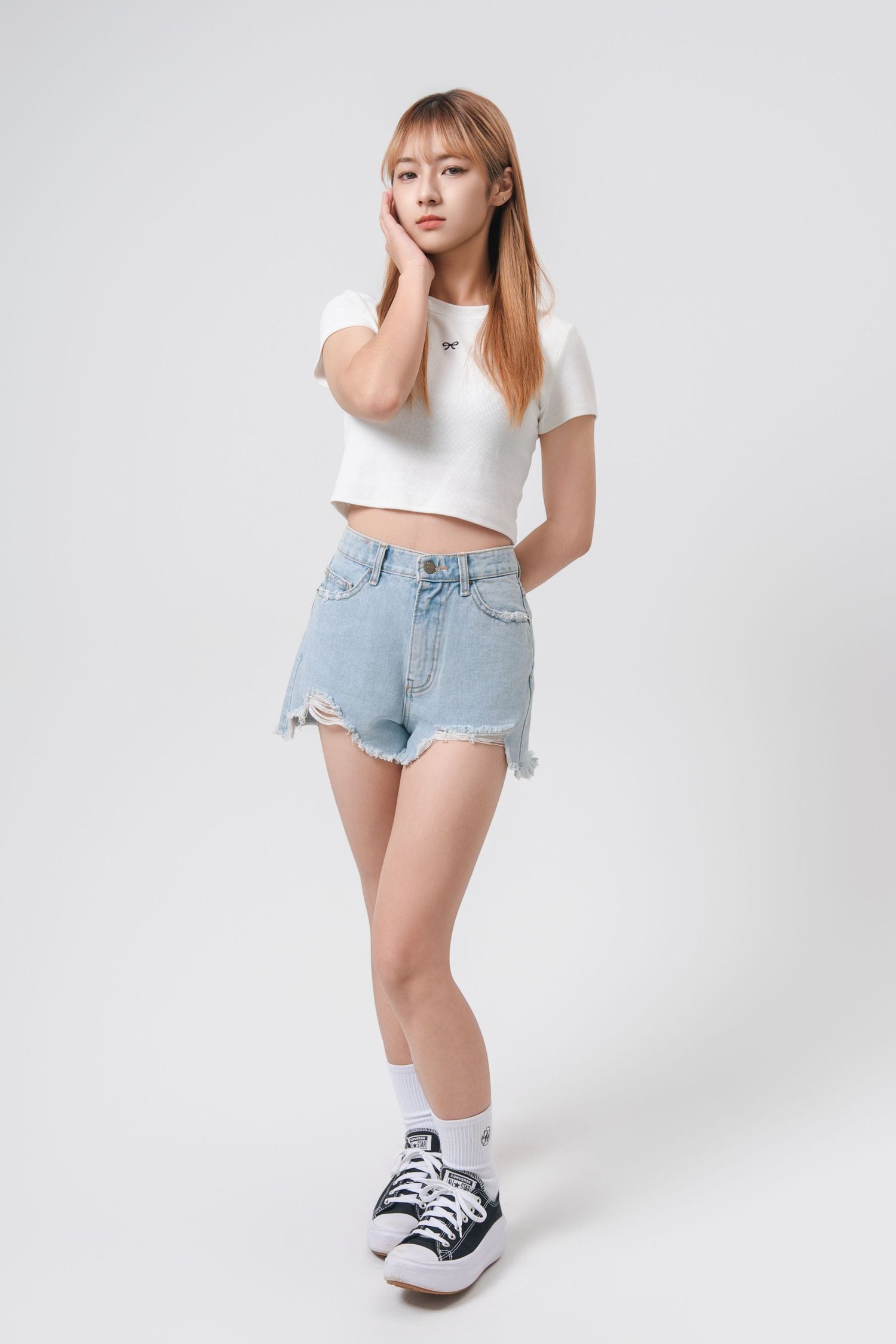 Person in casual white crop top and distressed denim shorts poses against white background with sneakers.