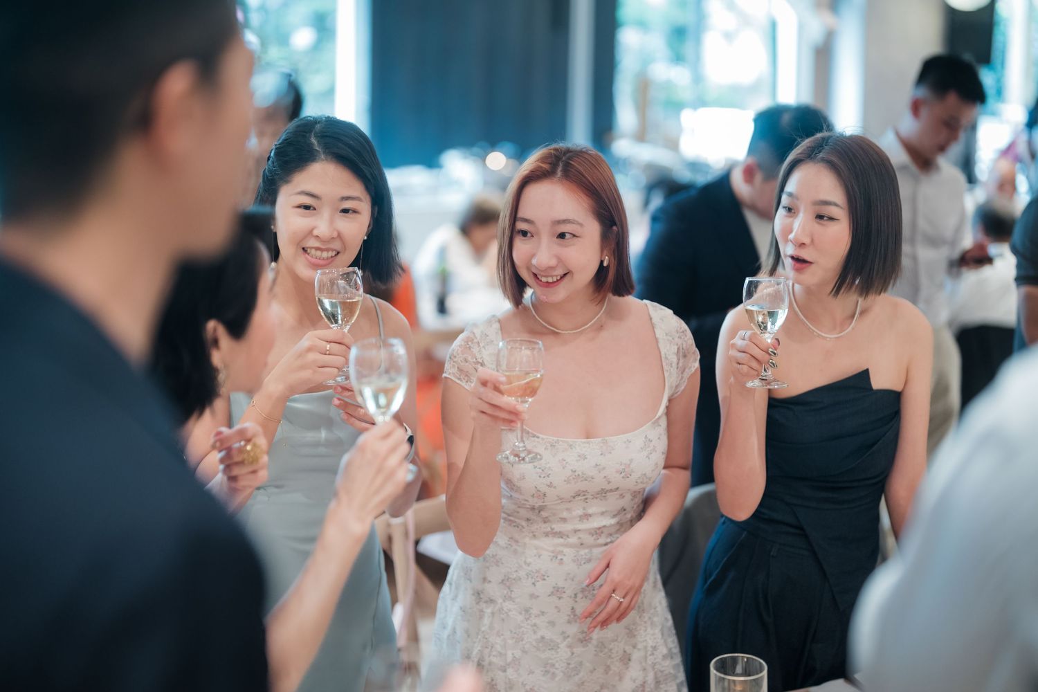 Wedding guests laugh and enjoy drinks at an elegant indoor reception in floral print and white dresses.