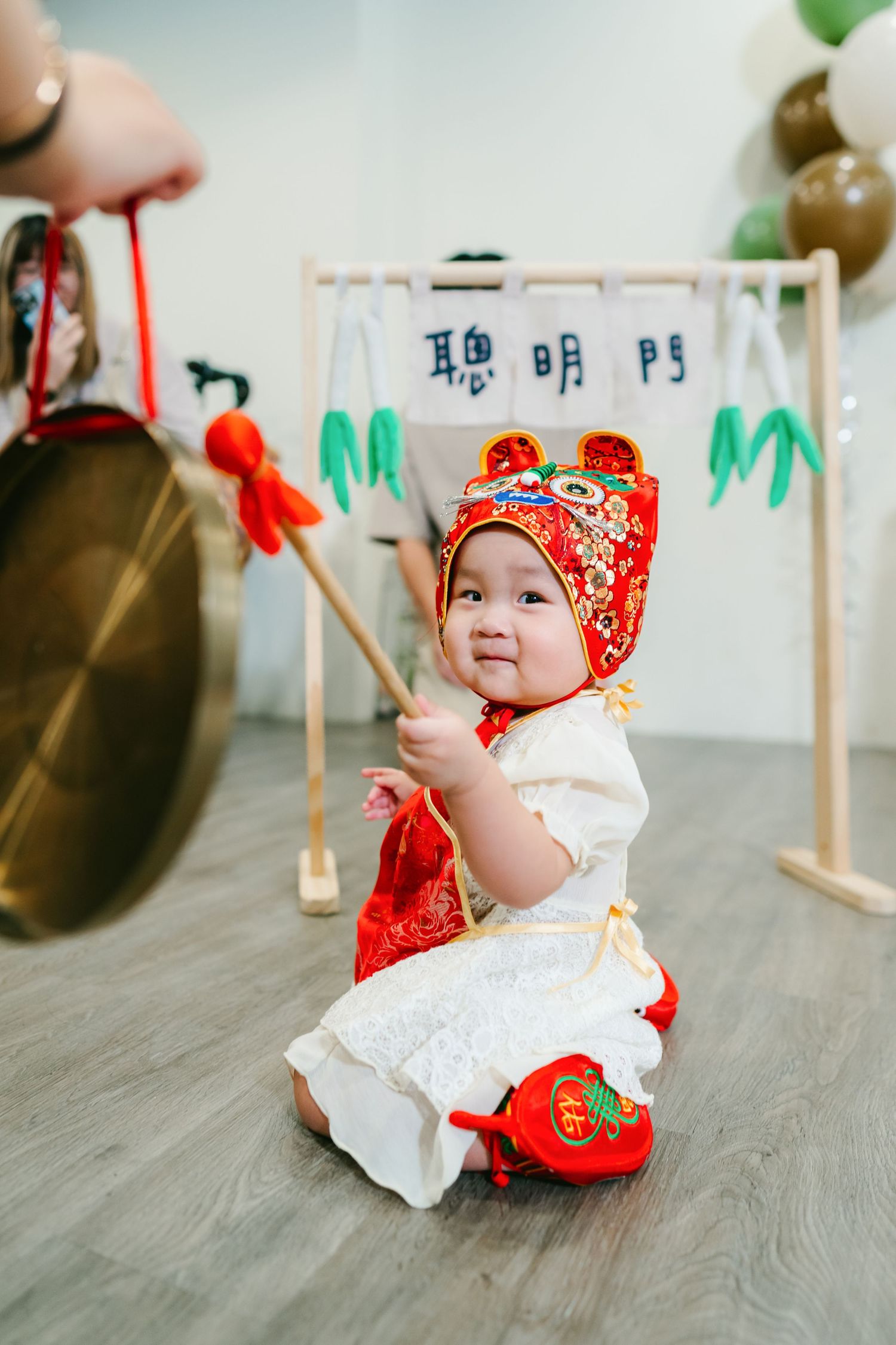 Small child wearing traditional red Chinese New Year outfit with matching hat sits on wooden floor holding a decorative stick.