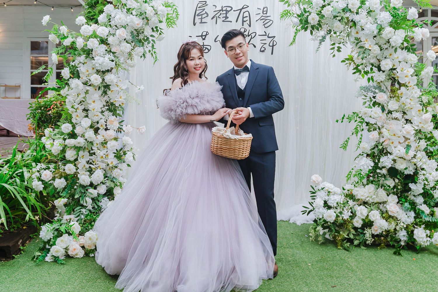 A romantic couple poses in a garden setting with lush white floral arrangements and green foliage.