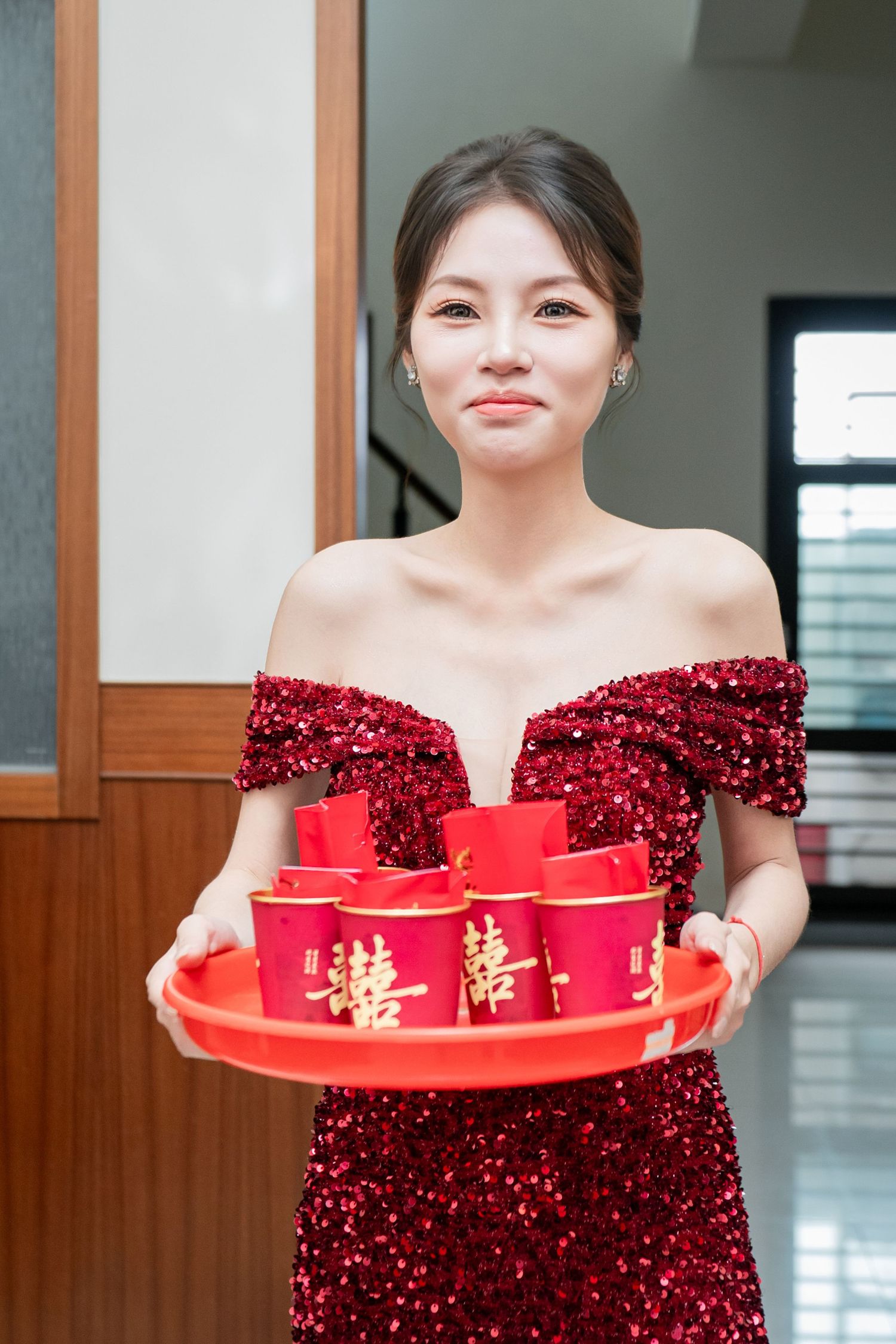 A person in a sparkly red off-shoulder dress holds a traditional Chinese tea serving tray with red cups.
