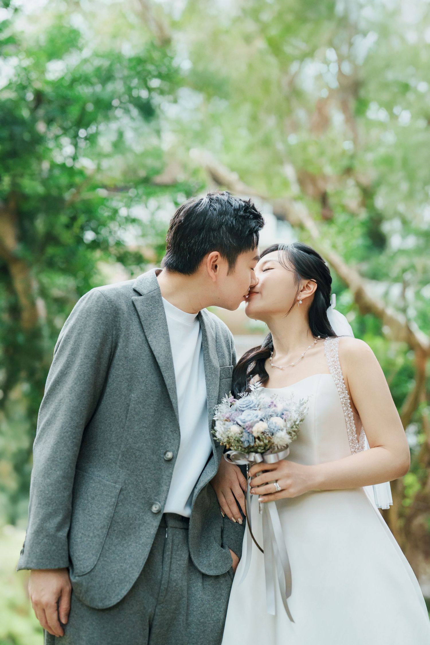 A romantic wedding couple shares tender kisses in a lush garden setting wearing gray suit and white dress.