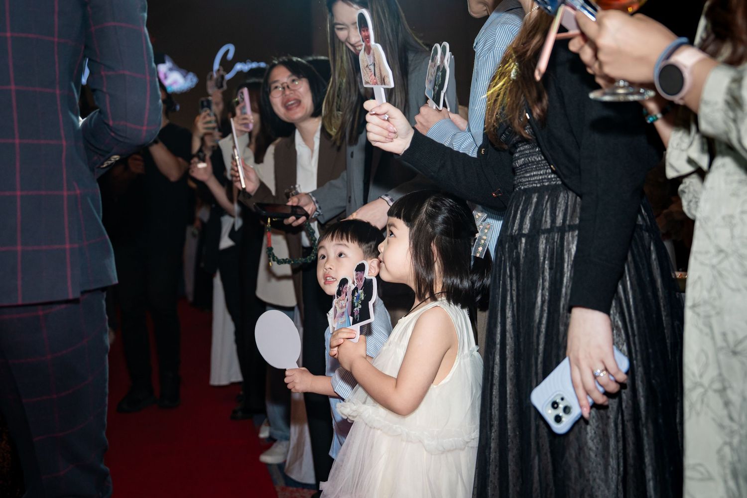 Young girl in white dress holds fan merchandise at a celebrity red carpet event surrounded by excited fans.