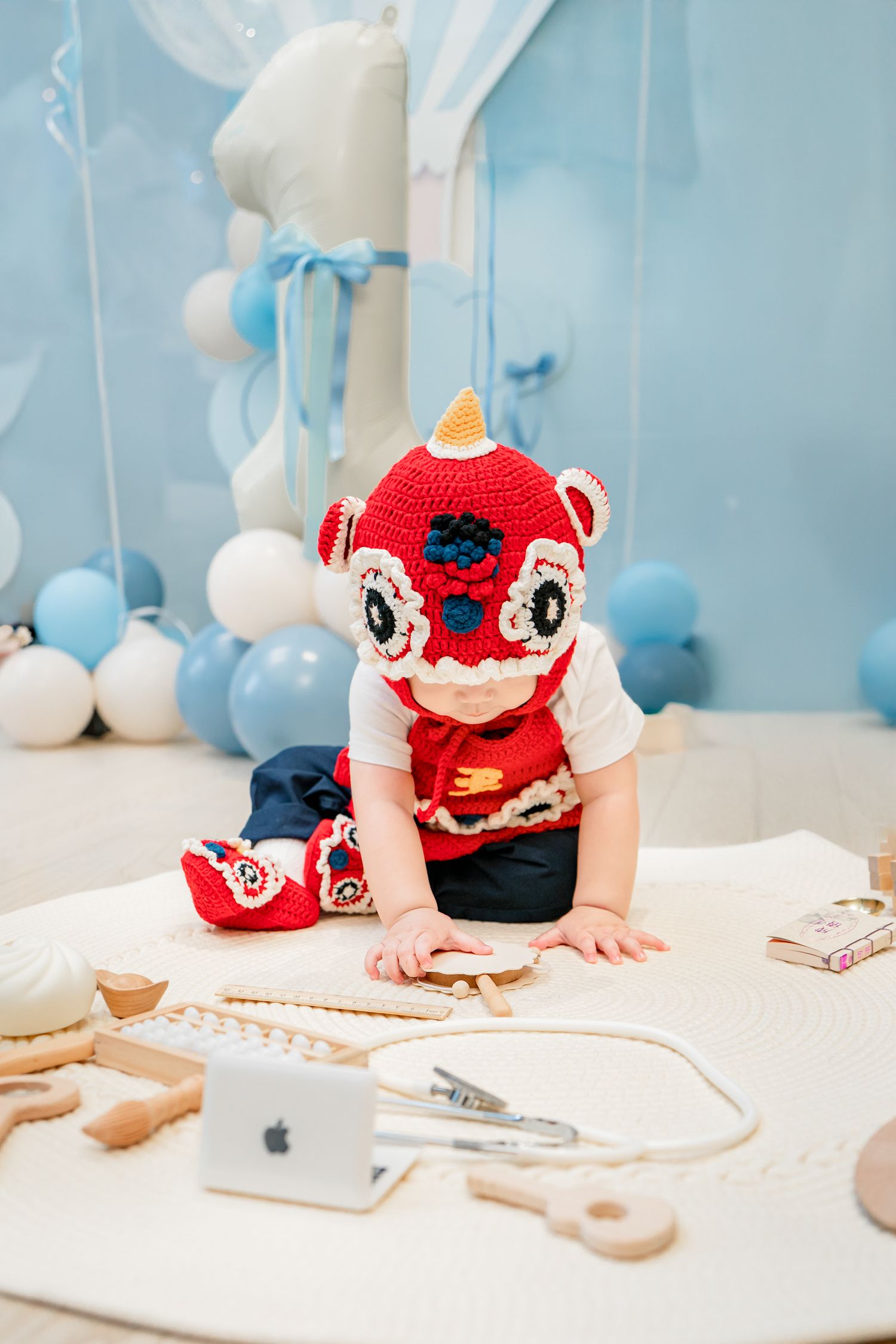 Child in red animal costume plays with toy medical instruments on white surface with blue balloons in background.