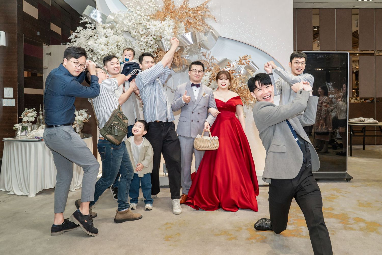 A celebratory group poses energetically at an indoor wedding reception with a person in a red gown as the centerpiece.