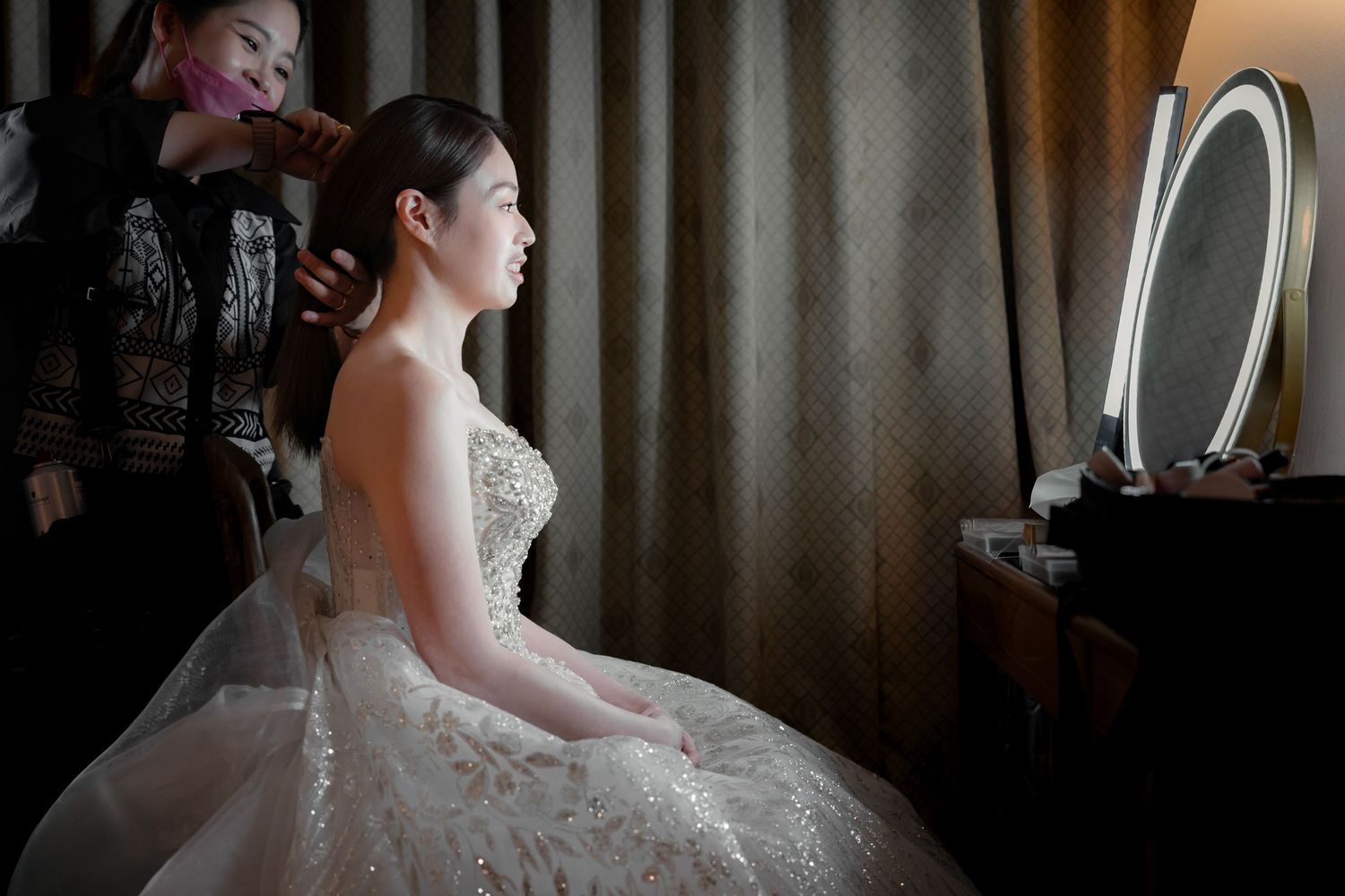 A bride in a white lace wedding dress gets ready in front of a mirror with soft lighting and elegant decor.