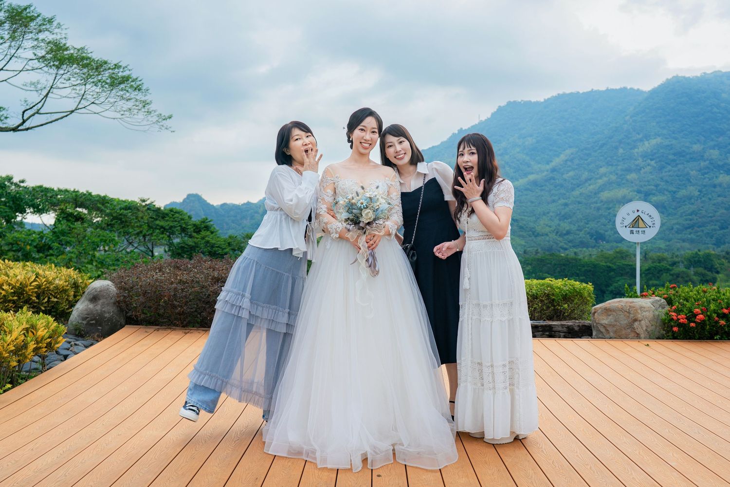 People in blue and white formal attire gather on a scenic wooden deck overlooking lush green mountains.