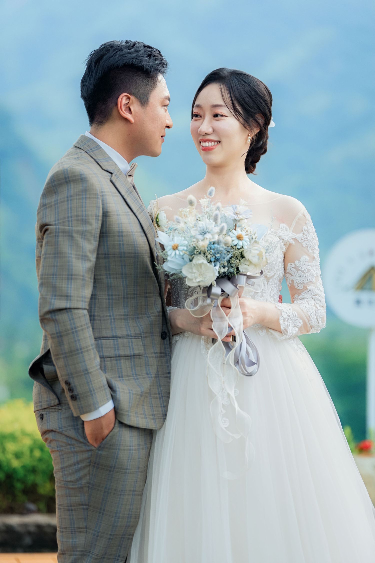 A couple in wedding attire pose together outdoors against a scenic mountain backdrop holding a white floral bouquet.