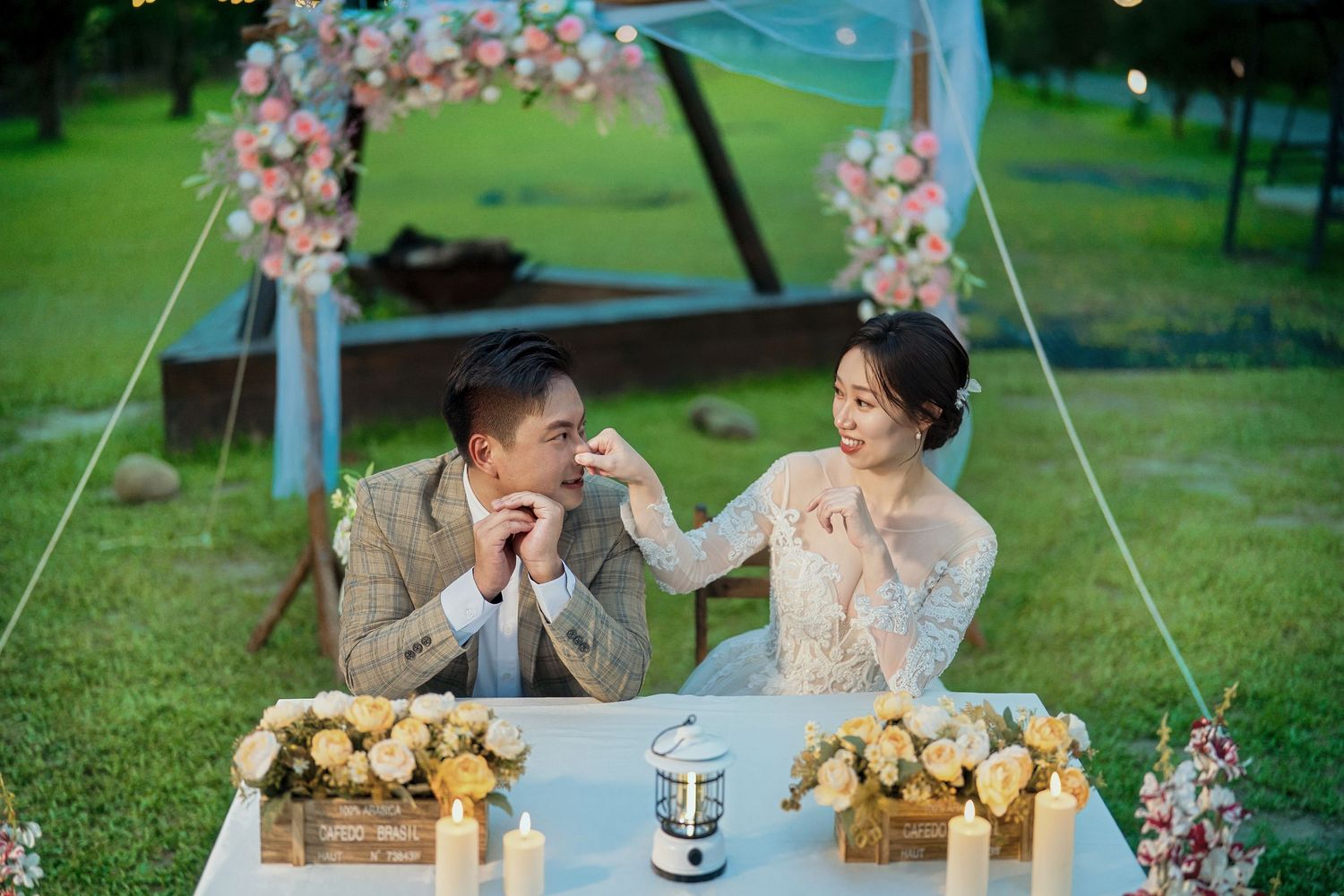 A romantic outdoor wedding scene with a couple seated at a decorated table under a floral arch in a garden setting.