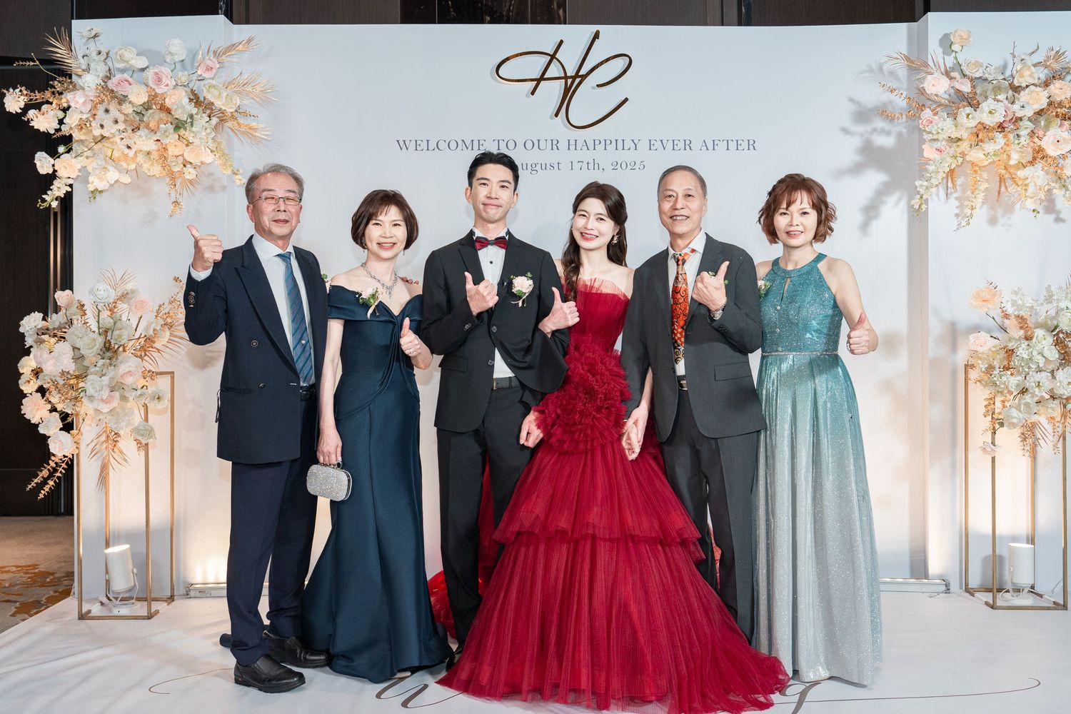 Wedding party stands together in formal attire with bride in striking red ball gown amidst elegant floral decor.
