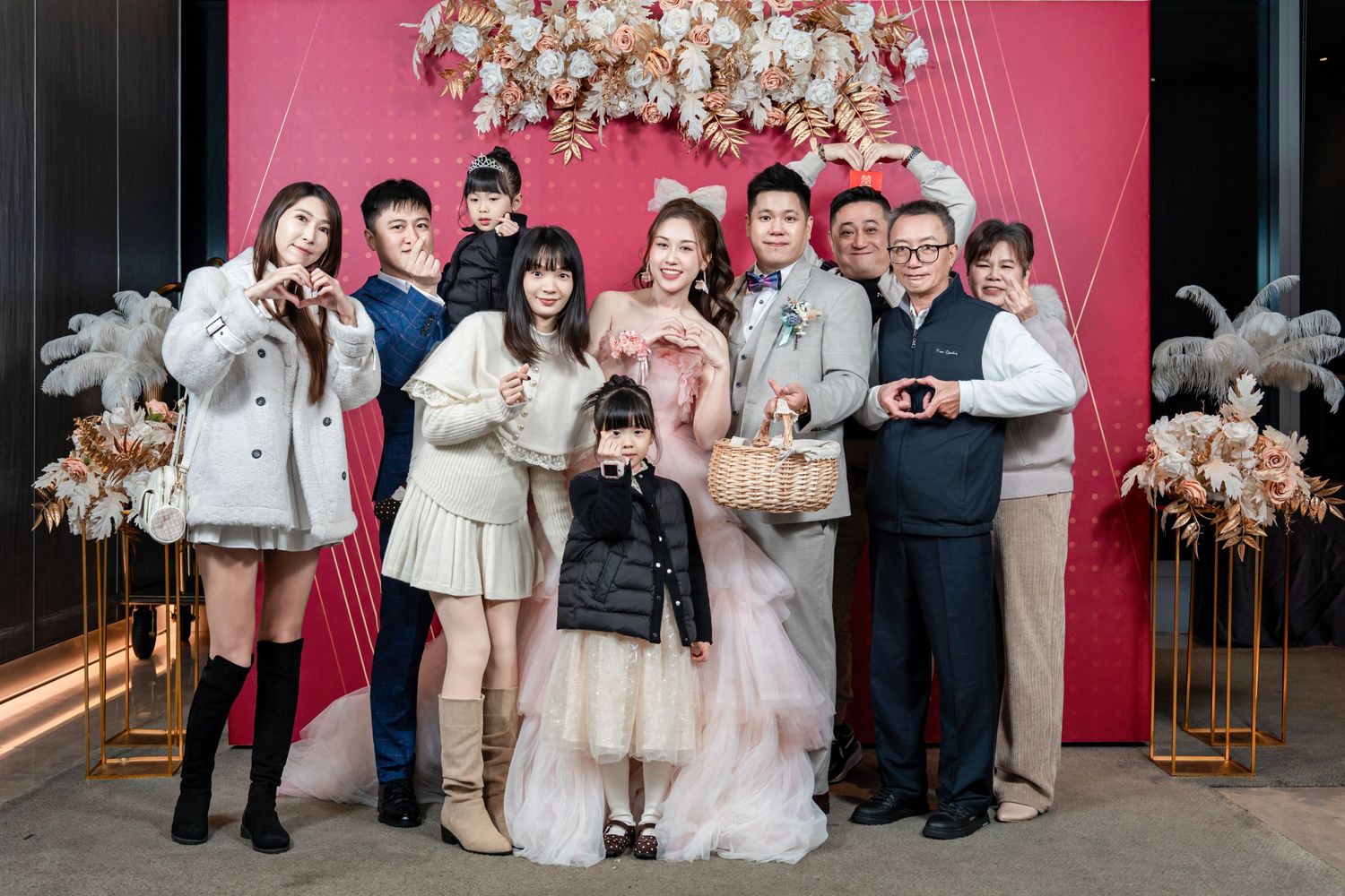 Wedding party members in formal attire pose together against a pink backdrop during an indoor celebration.