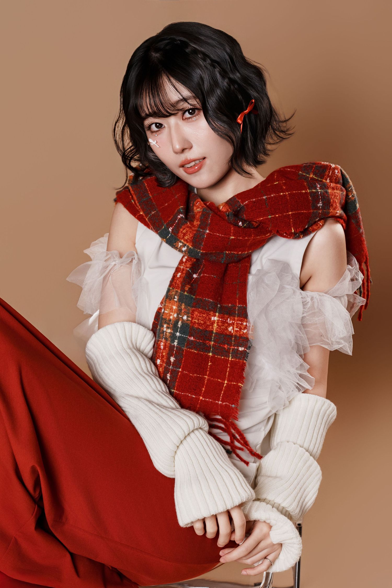 A model wearing a red plaid scarf and white ruffled sweater poses against a warm brown background in a fashion photoshoot.