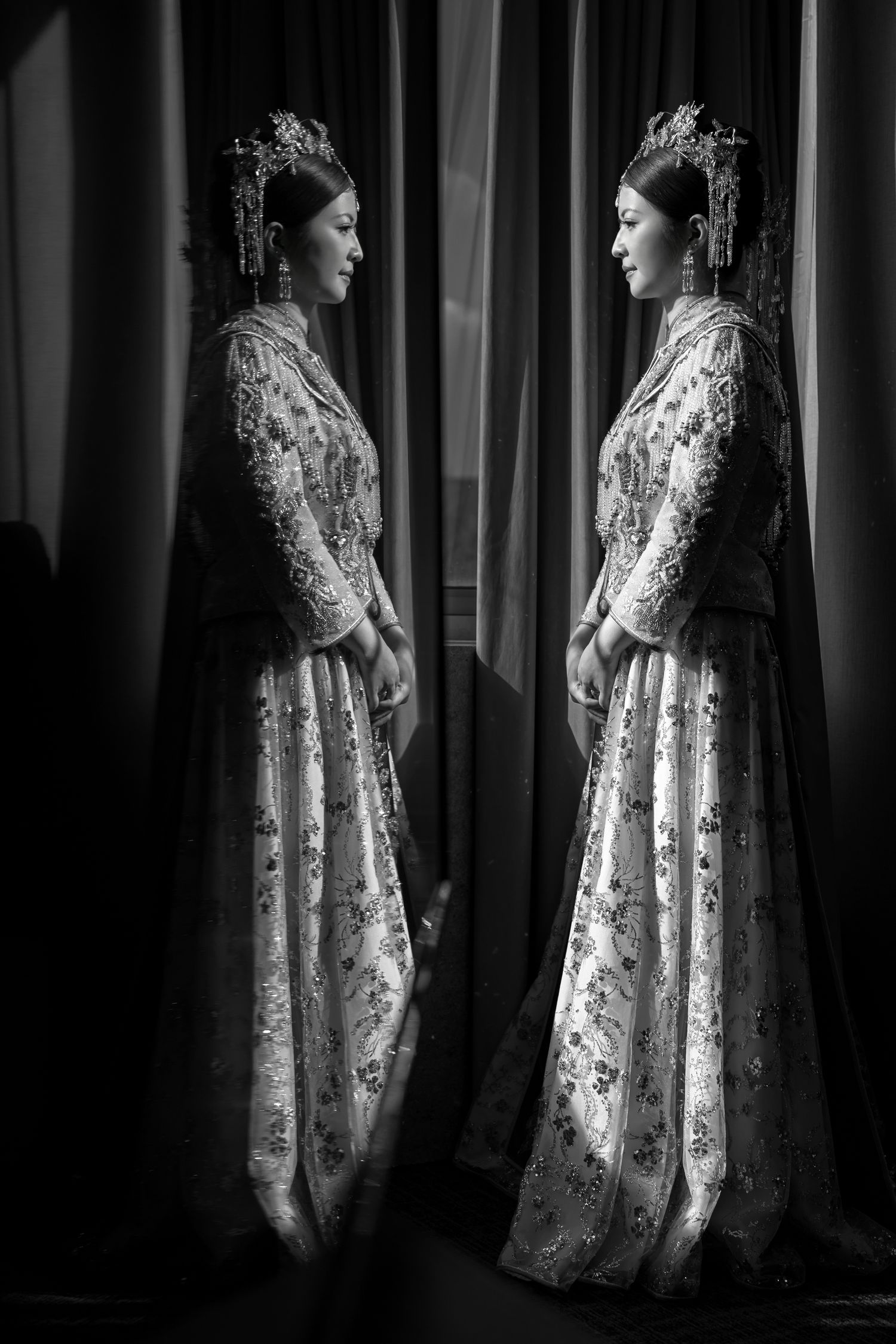 Black and white photo of a bride in an ornate embroidered gown and headdress standing beside a mirror, her reflection facing her.