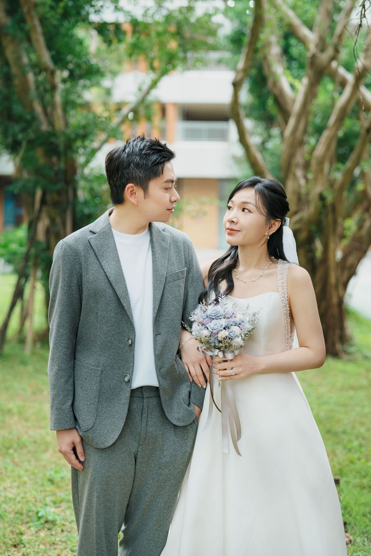 A couple in wedding attire stands together in a garden setting with trees in the background.