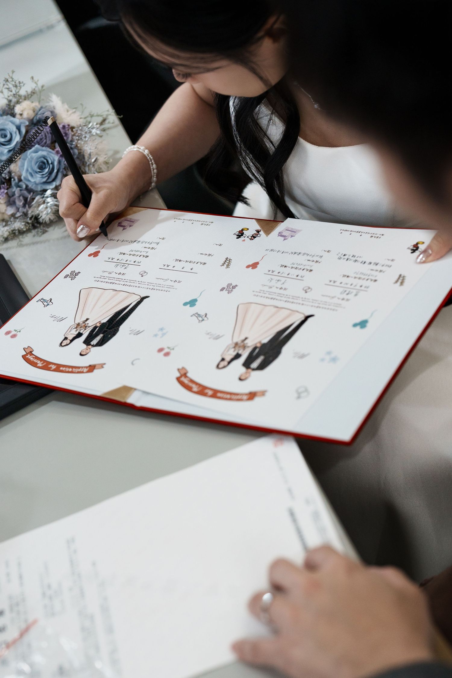 Someone writing in a guest book or signing document with illustrated wedding couple designs on the pages.