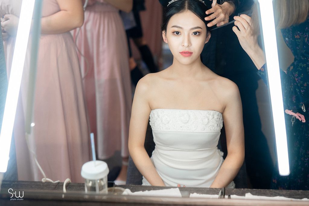 A bride in a strapless gown sits at a vanity with bright lighting during wedding preparations.