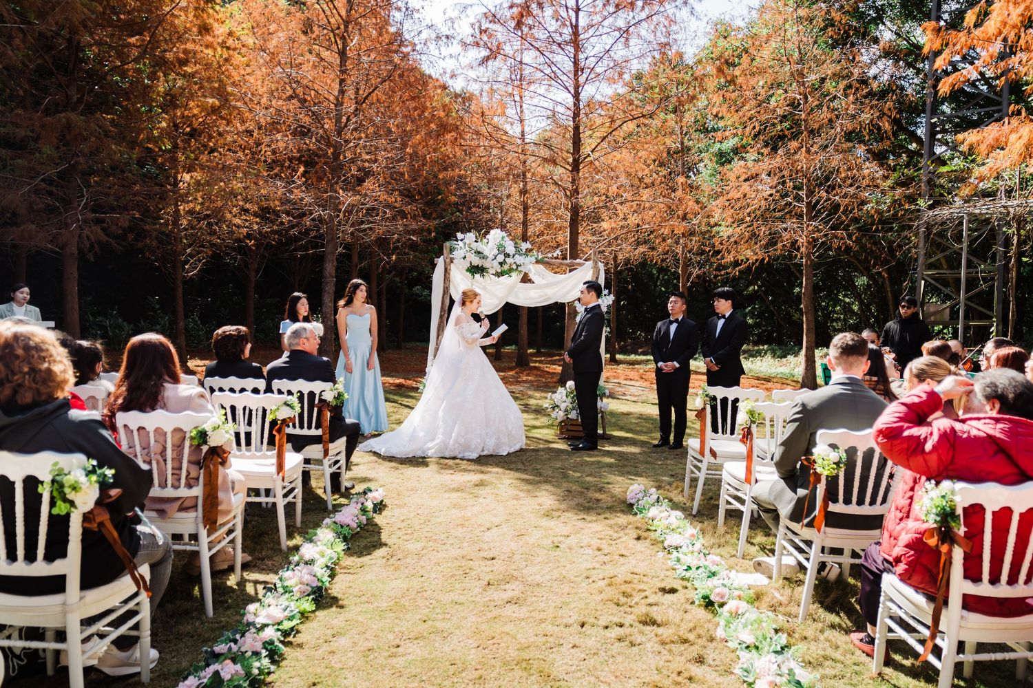 Outdoor autumn wedding ceremony with white chairs and floral-lined aisle under golden foliage.