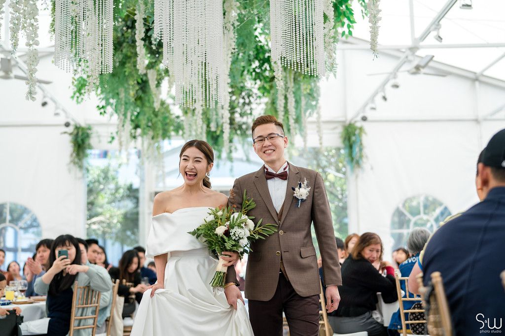 A couple walks down the aisle in wedding attire under hanging greenery decor.
