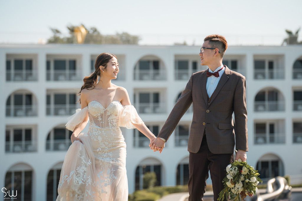 A wedding couple poses in front of a white Mediterranean-style resort building on a sunny day.