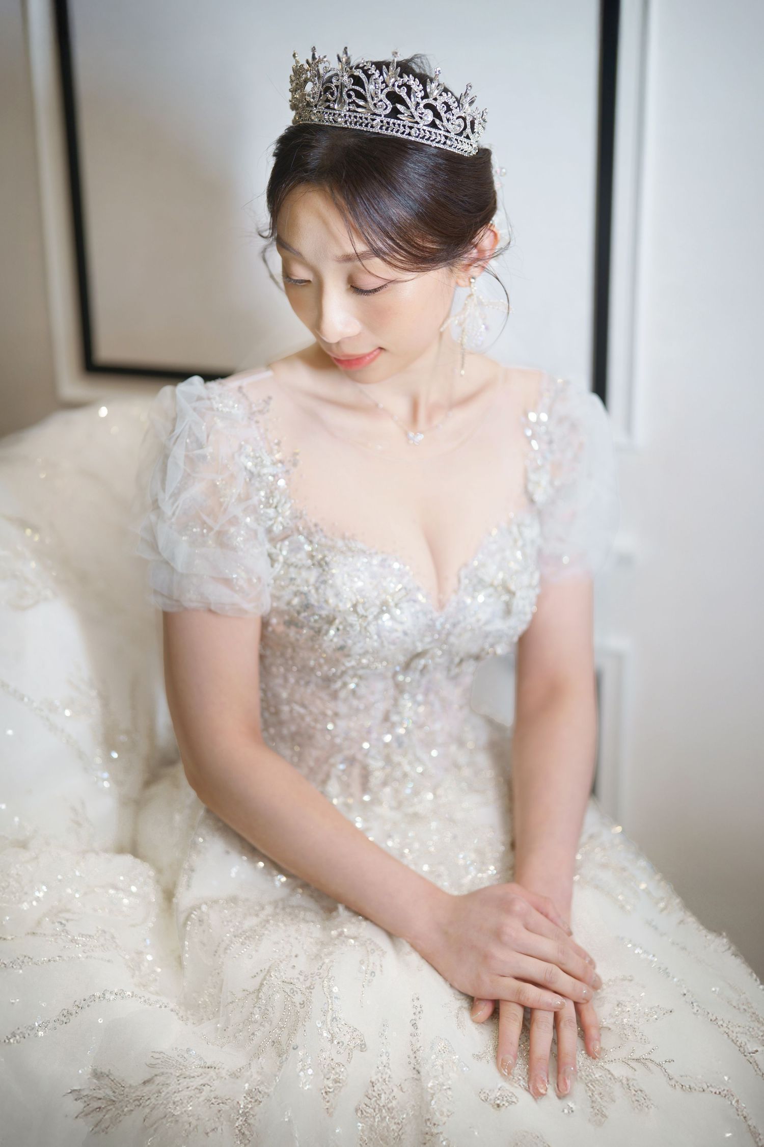 A bride wearing a sparkling white wedding gown with short sleeves and a crystal tiara sits elegantly in soft lighting.