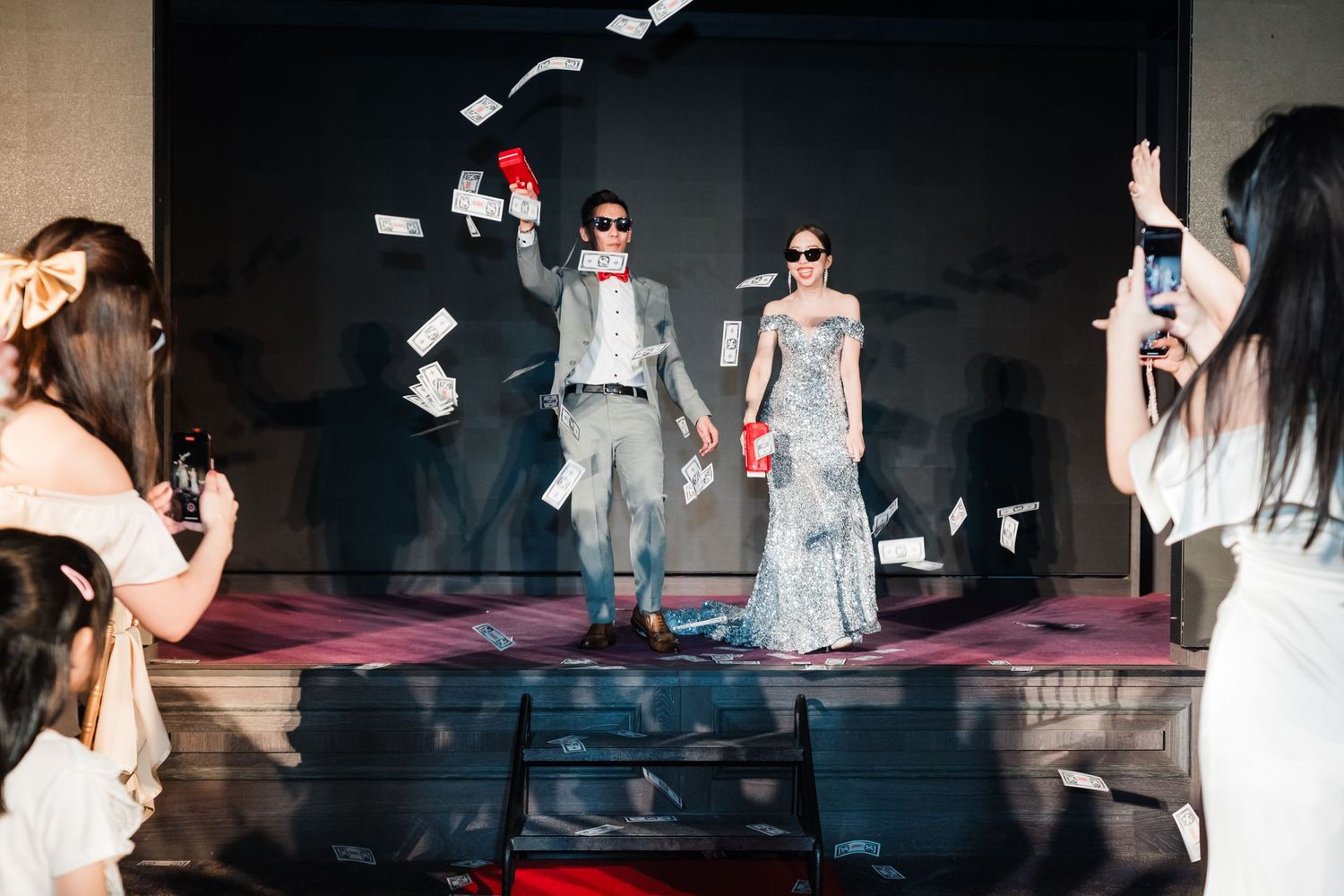 Wedding celebration on stage with money being thrown in the air as couple stands on dark platform with photographers.