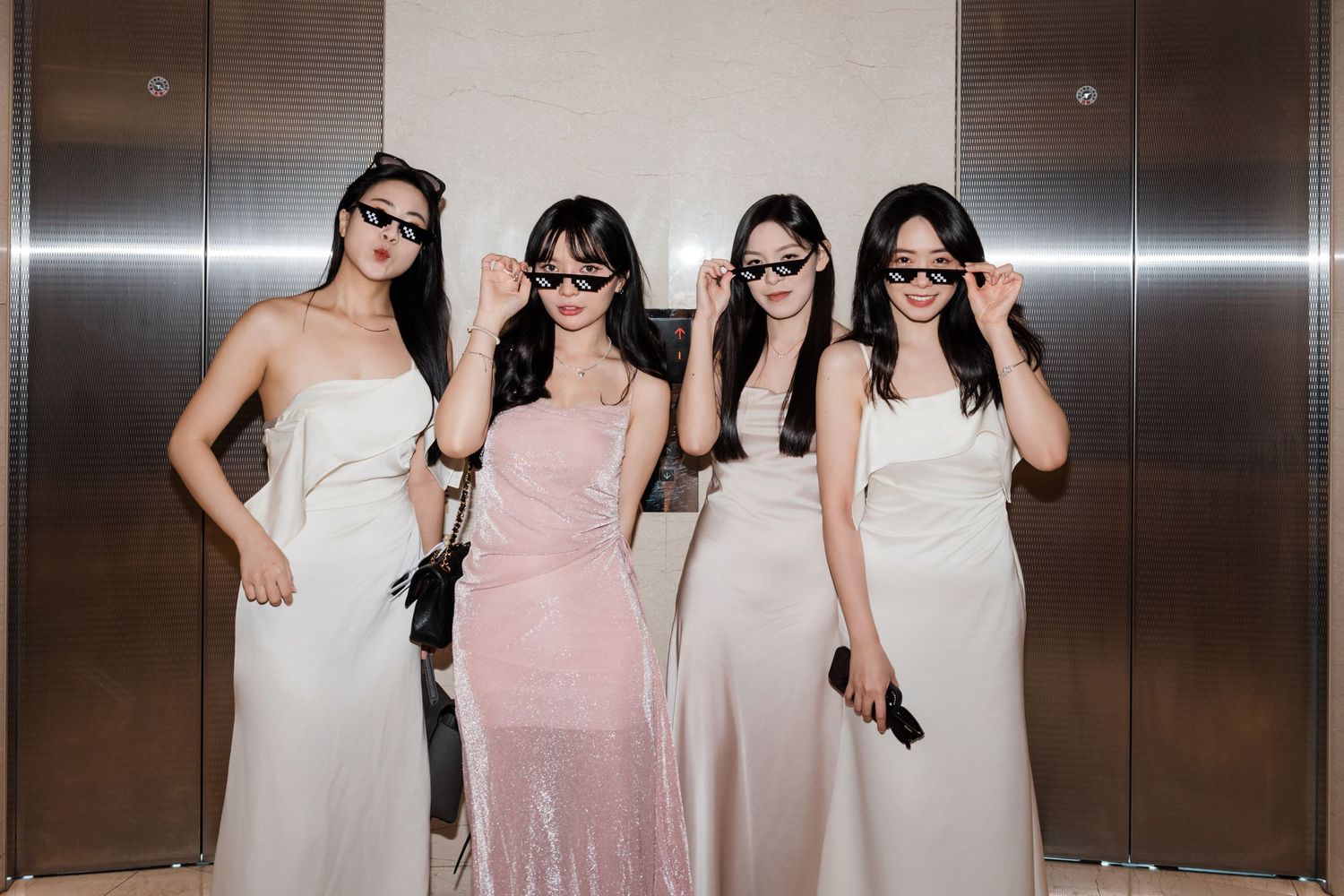 Four women in elegant evening gowns pose together while wearing sunglasses in front of an elevator.