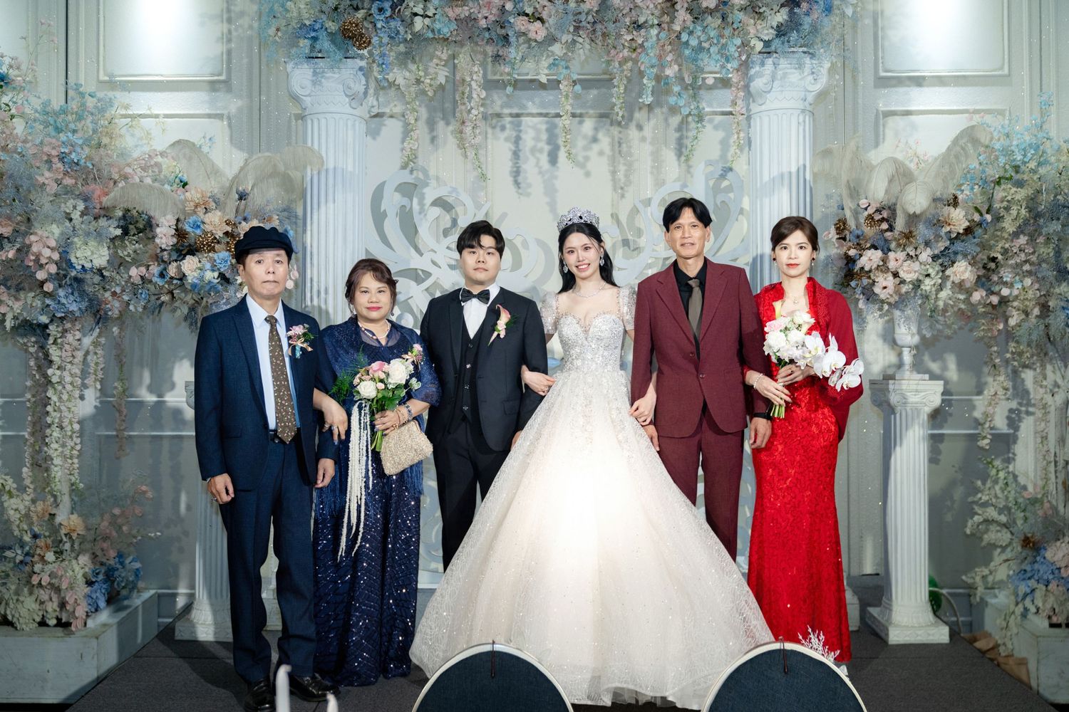 A formal wedding celebration group photo with floral decorations and elegant attire against an ornate grey backdrop.