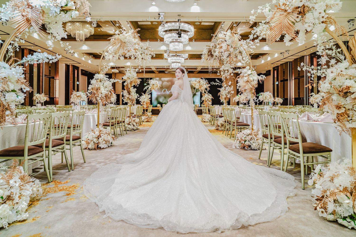 Luxurious wedding reception venue decorated with ornate floral arrangements and crystal chandeliers shows a flowing white gown.