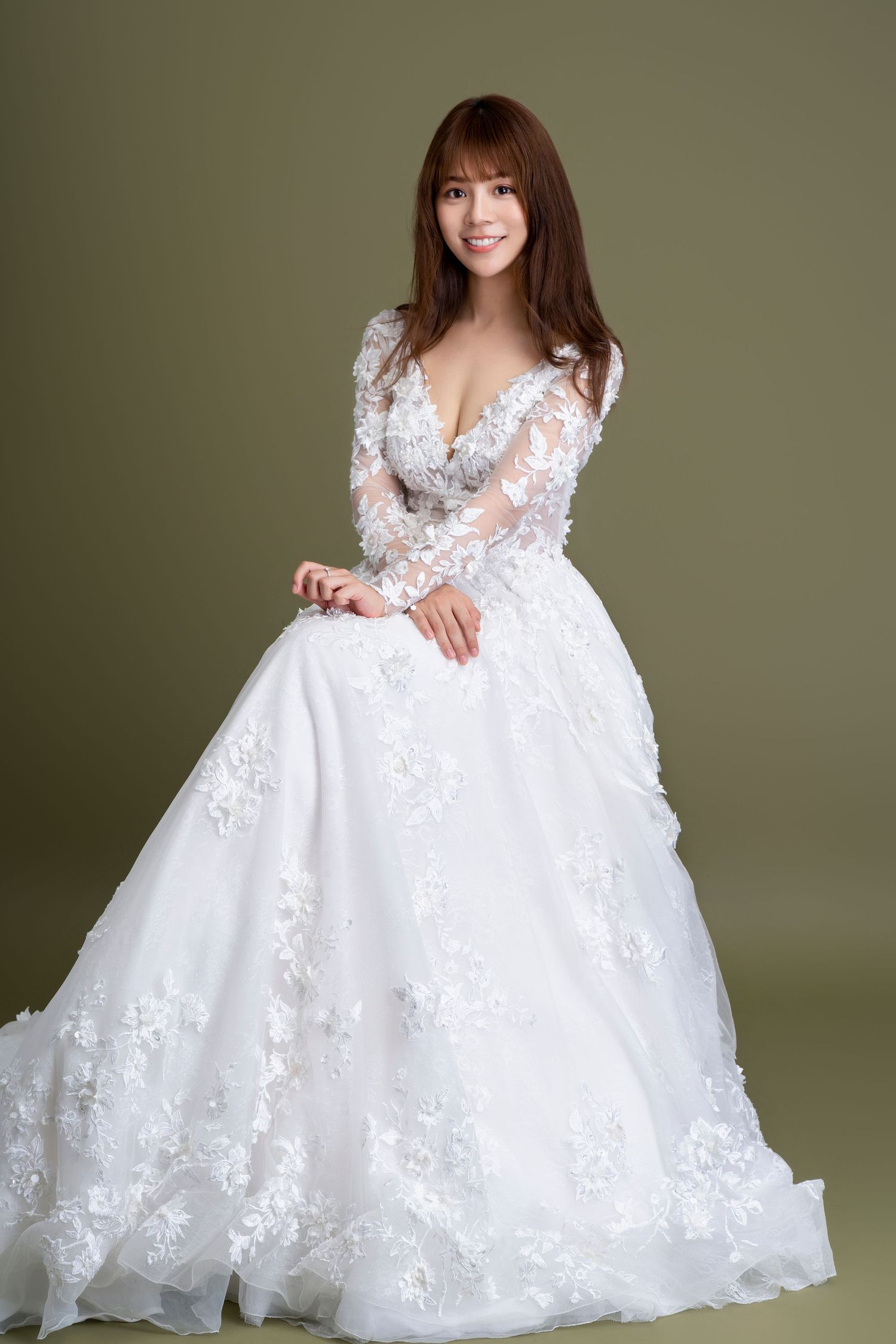 A wedding dress with long lace sleeves and floral appliques on a flowy white ballgown silhouette against olive background.