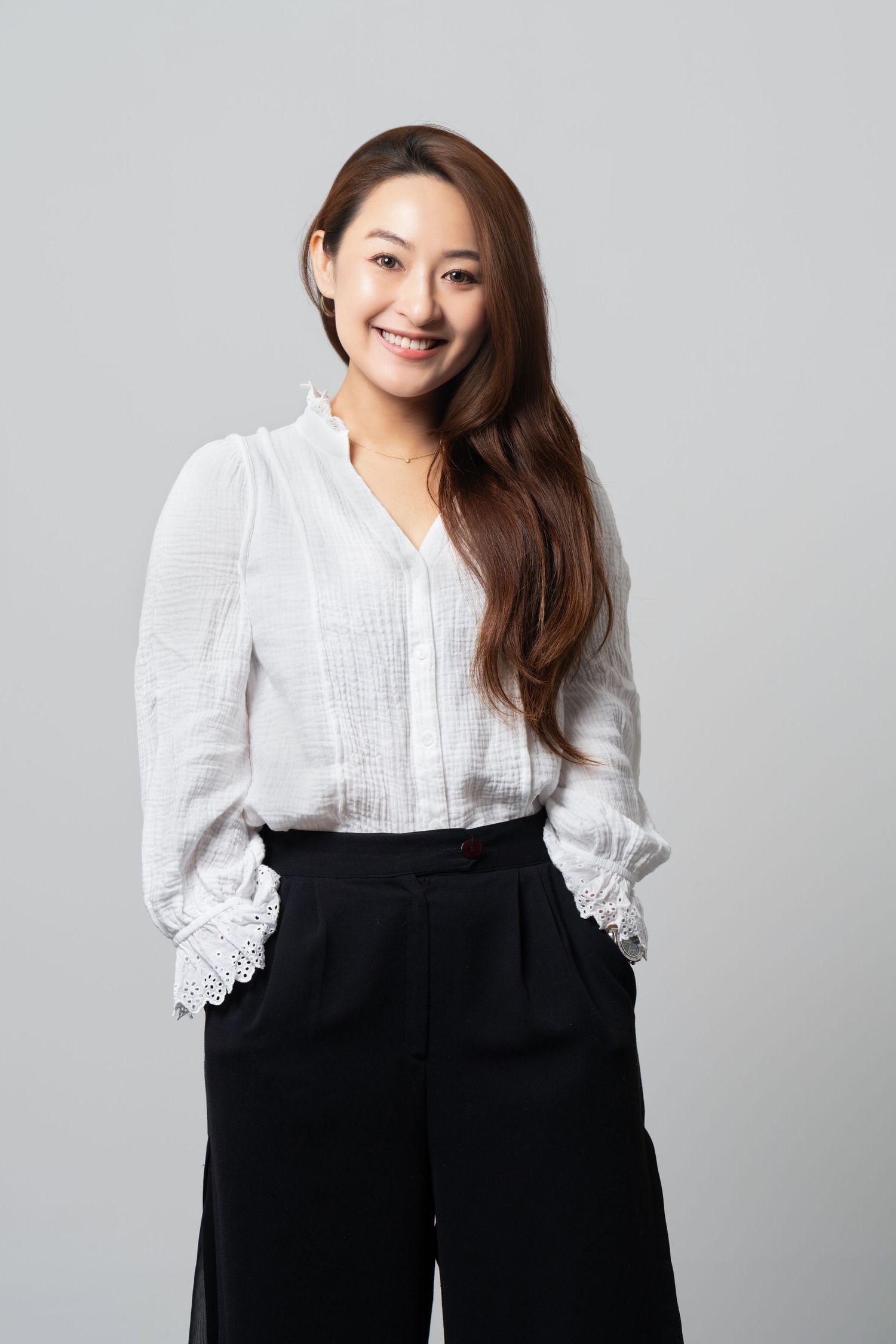 Professional portrait of a person in a white lace-trimmed blouse and black wide-leg pants against a gray background.