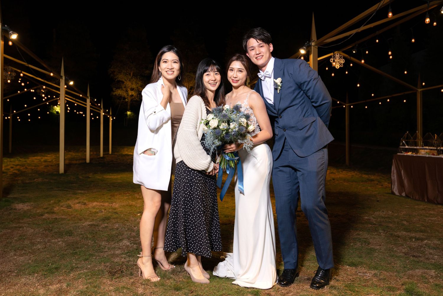 People pose together at an outdoor evening wedding reception with string lights overhead.