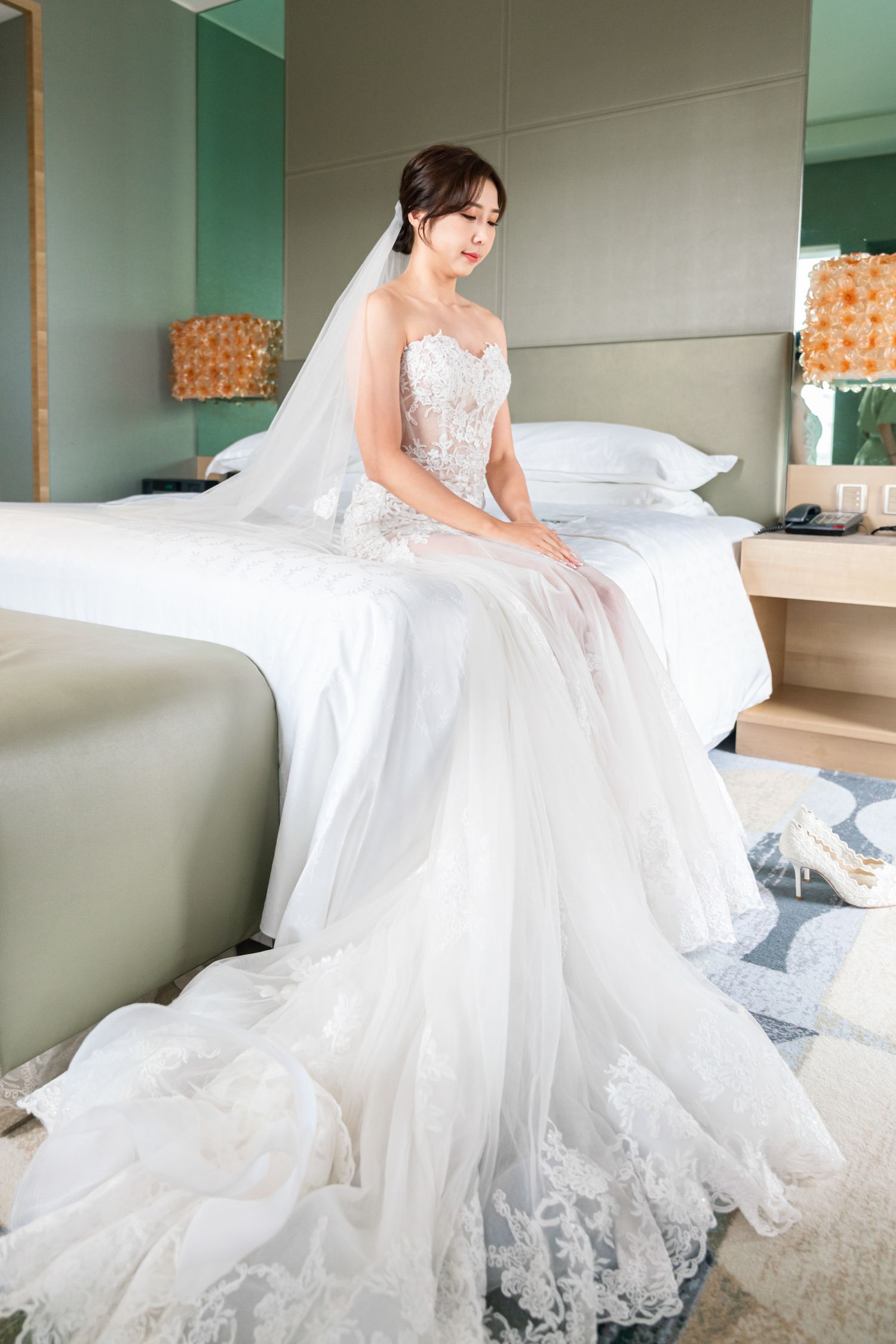 A bride in an elegant white lace wedding gown sits gracefully on a hotel bed, with her veil flowing behind her.