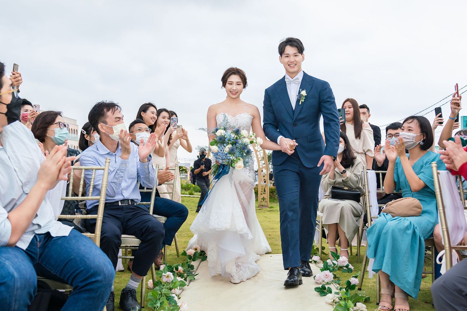 Wedding guests sitting in white chairs celebrate during an outdoor ceremony along a grassy aisle decorated with greenery.