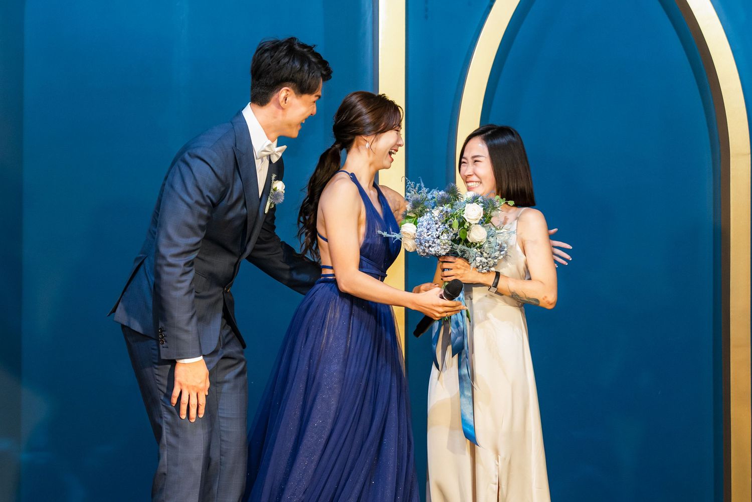 Three people in formal attire stand against a blue wall with an arched gold frame, sharing a joyful moment with flowers.