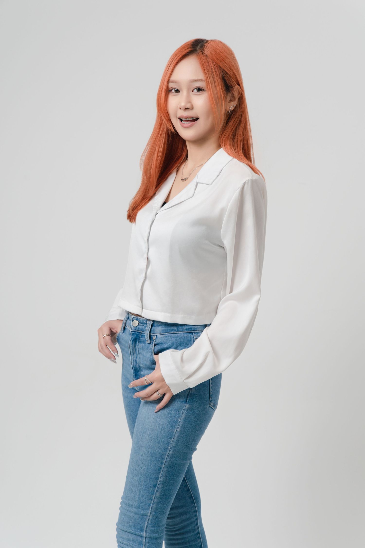 Woman in white long sleeve blouse and high-waisted blue jeans poses against light background in studio photoshoot.