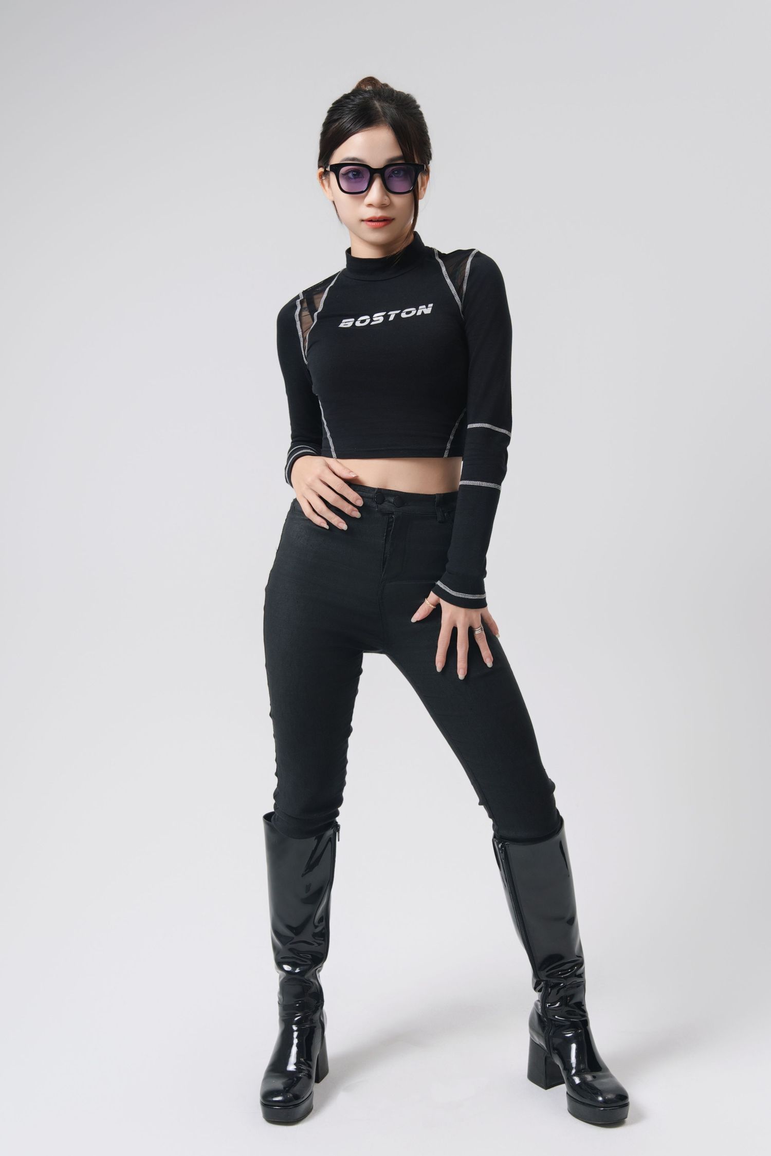 Model in black crop top, high-waisted pants and chunky boots poses against white background wearing sunglasses.